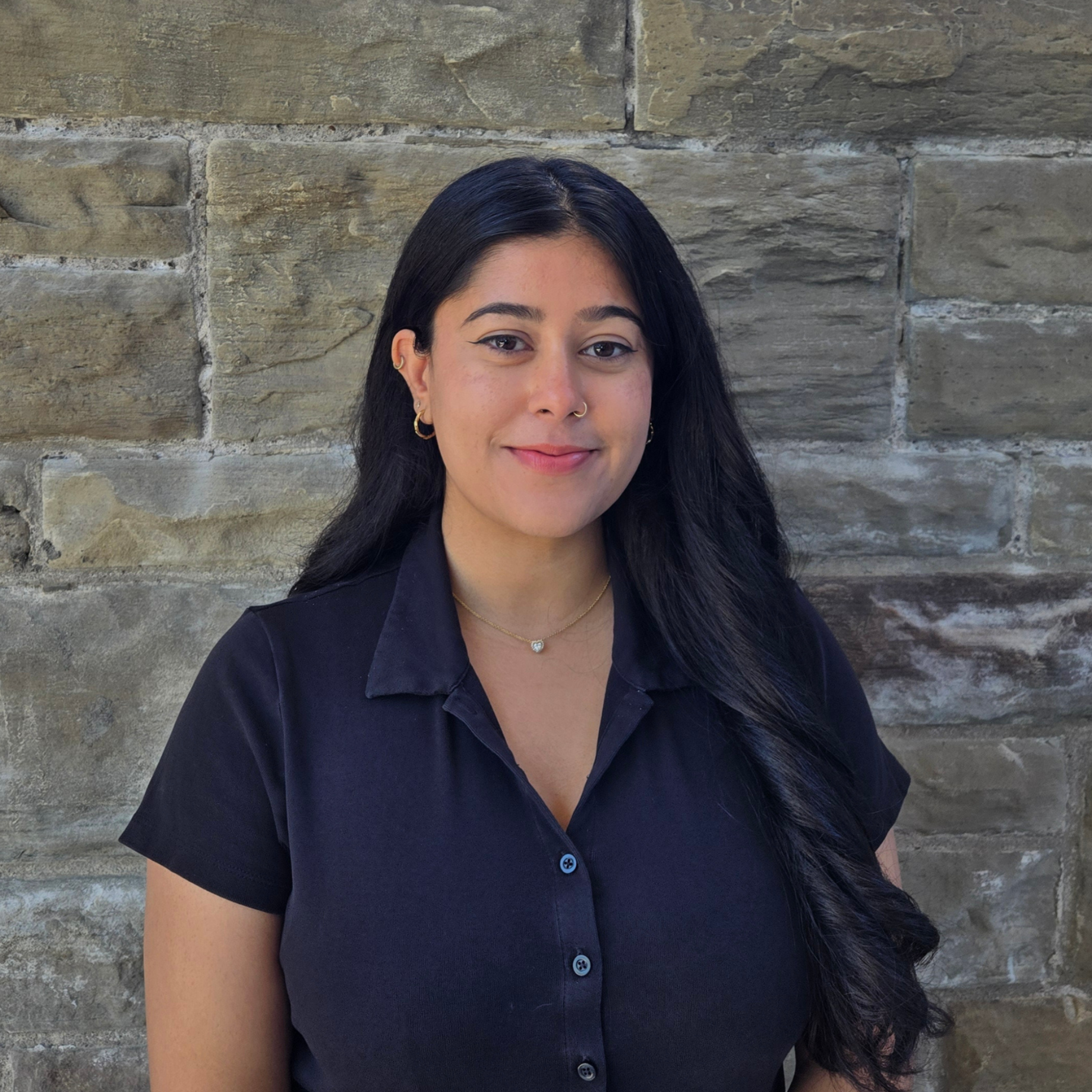 A young woman sitting on a chair against a textured beige stone wall. She has long black hair, is smiling slightly, and is wearing a brown plaid blazer over a white top. She has small hoop earrings, a nose ring, and a necklace with a heart-shaped pendant.