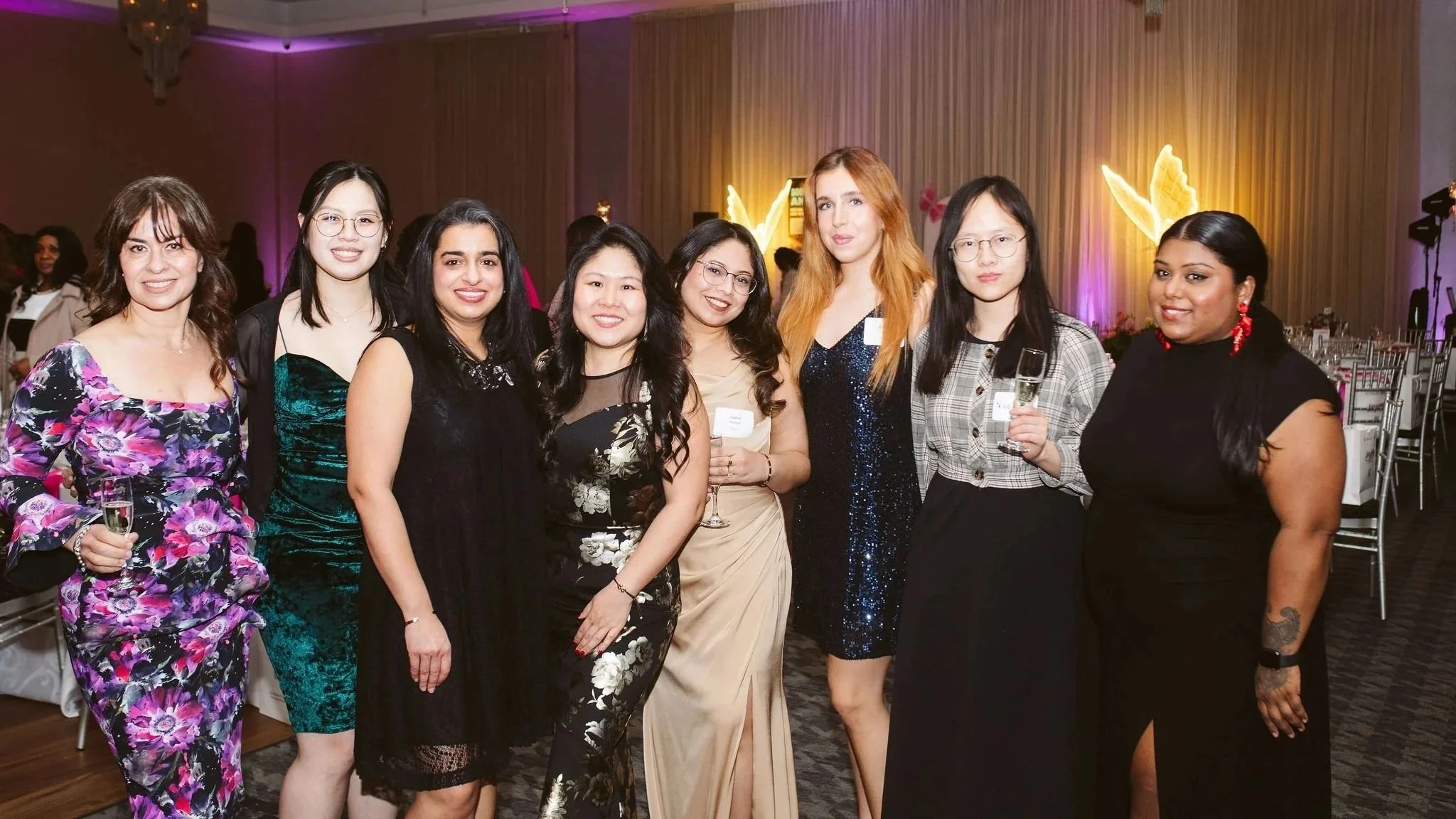Group of nine women dressed in formal attire at an indoor event, some holding glasses of champagne, with purple lighting and angel wing decorations in the background.