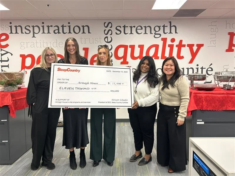 Group of five women standing in an office, holding a large check for $11,000 made out to Armagh House, smiling, with a decorated table and wall in the background featuring words like 'quality', 'strength', and 'inspiration'.