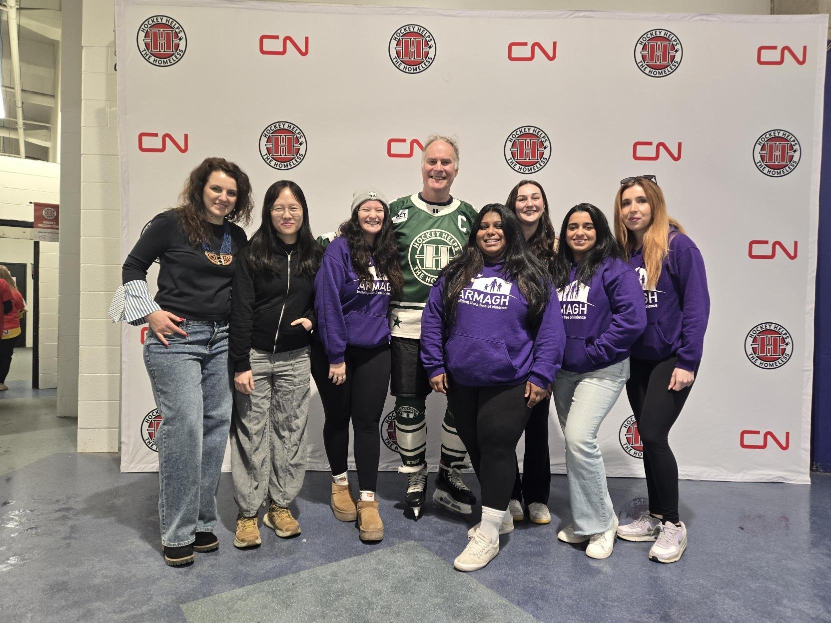 Group of 9 people, including a man wearing hockey gear, standing in front of a backdrop with logos. The people are smiling, and some are wearing purple hoodies with the word 'ARMAGH' and an image of a family holding hands.