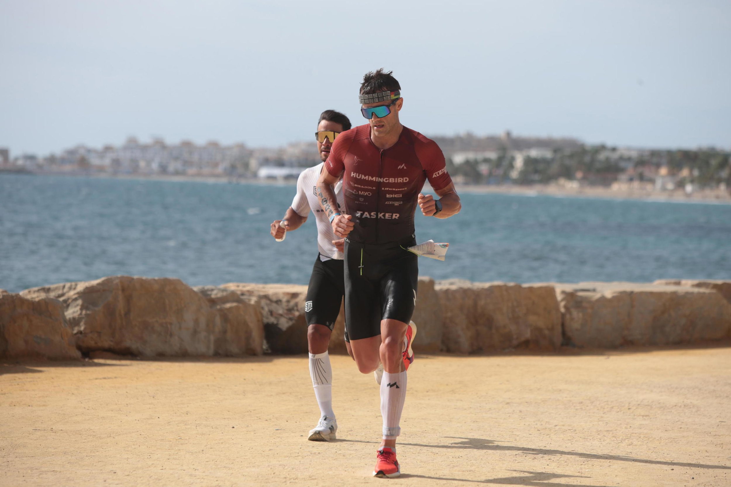 Two male athletes running along a coastal path with the ocean and city in the background, wearing sports gear, sunglasses, and headbands.