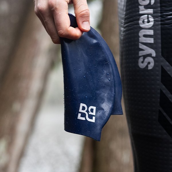 Person holding a wet blue swim cap with a logo, wearing a wetsuit outdoors.