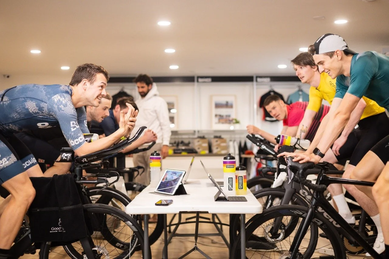 Group of cyclists in indoor training session, riding stationary bikes, engaging in a discussion or coaching, with tablets and water bottles on a table in front of them.