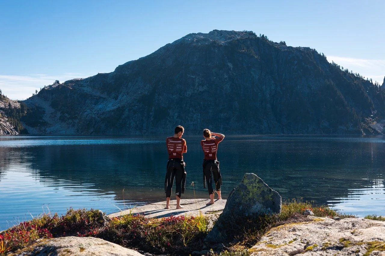 Two people standing on a rocky shoreline looking at a mountain across a calm lake, with clear blue sky overhead.