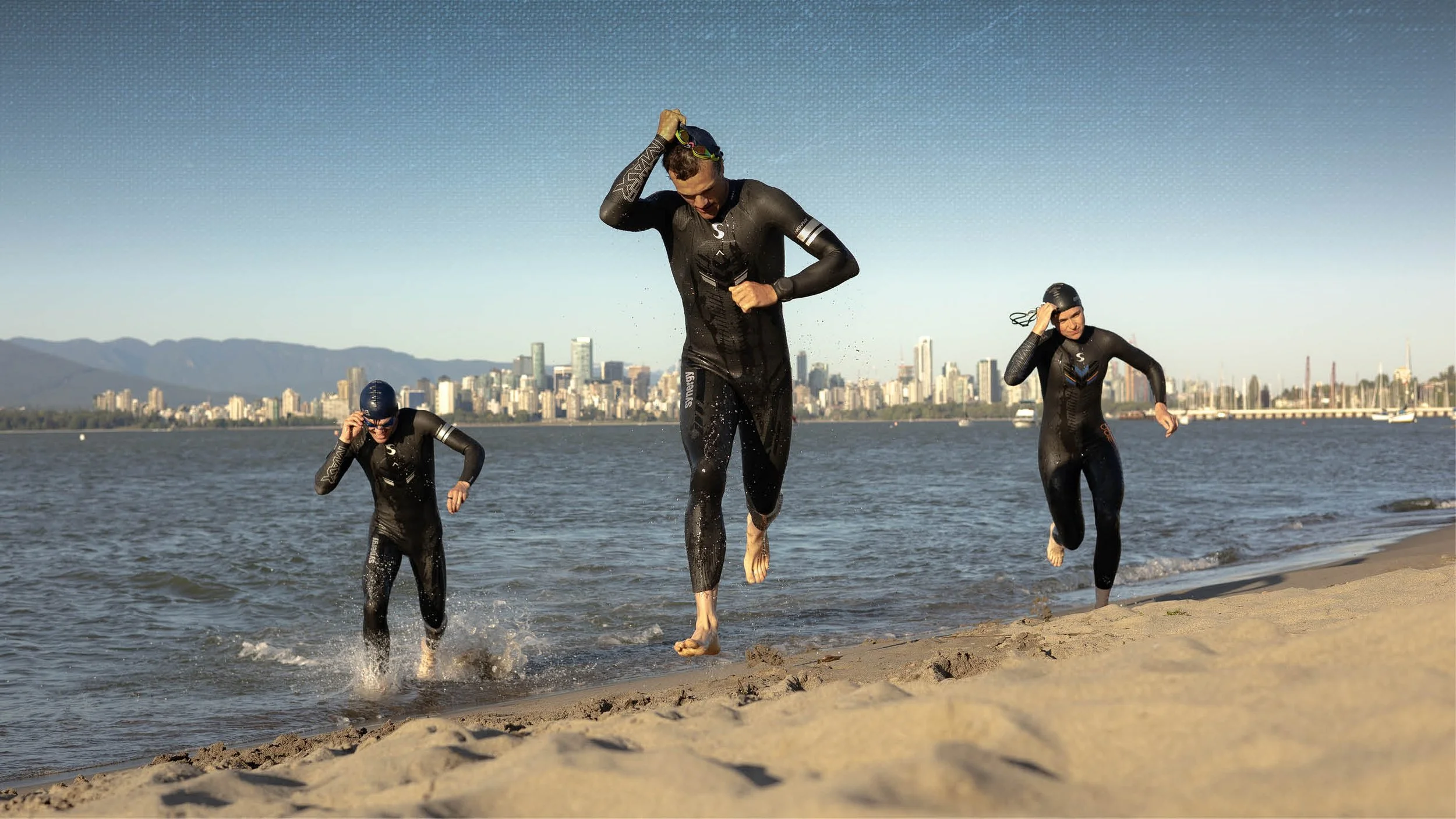 Three athletes in wetsuits emerging from the water onto a sandy beach as a city skyline and mountains are visible in the background.