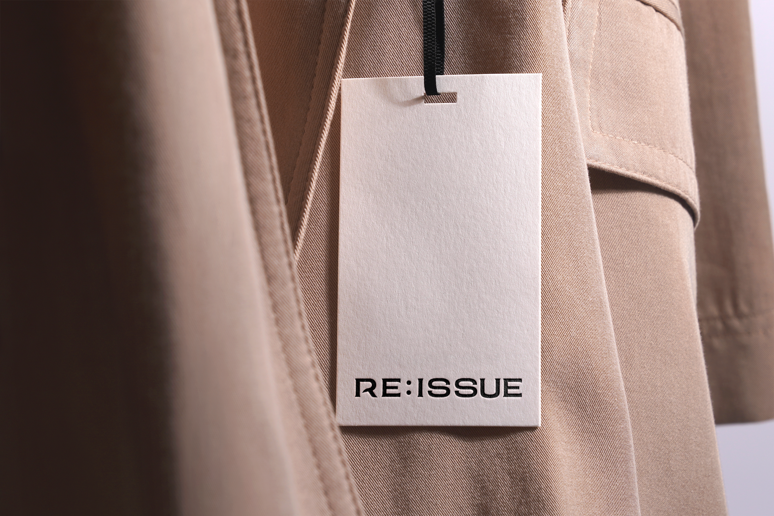 Close-up of a beige trench coat with a white tag attached that reads 'RE: ISSUE' in black text.