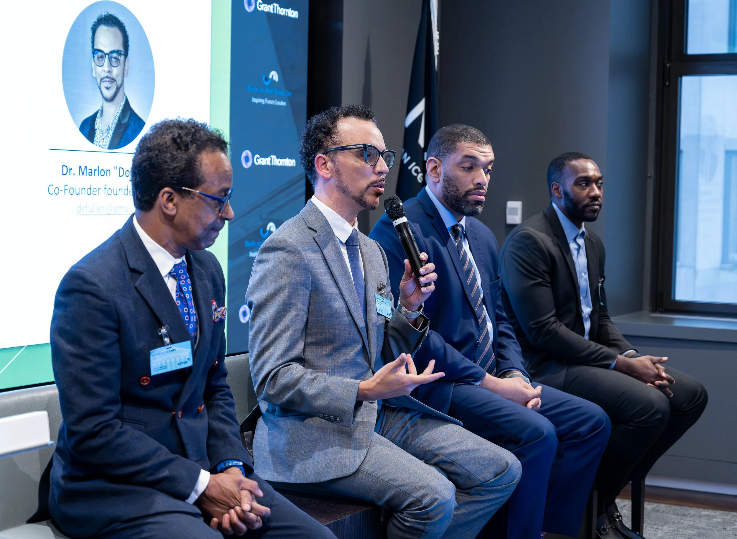 Four men in suits participating in a panel discussion, with one holding a microphone.