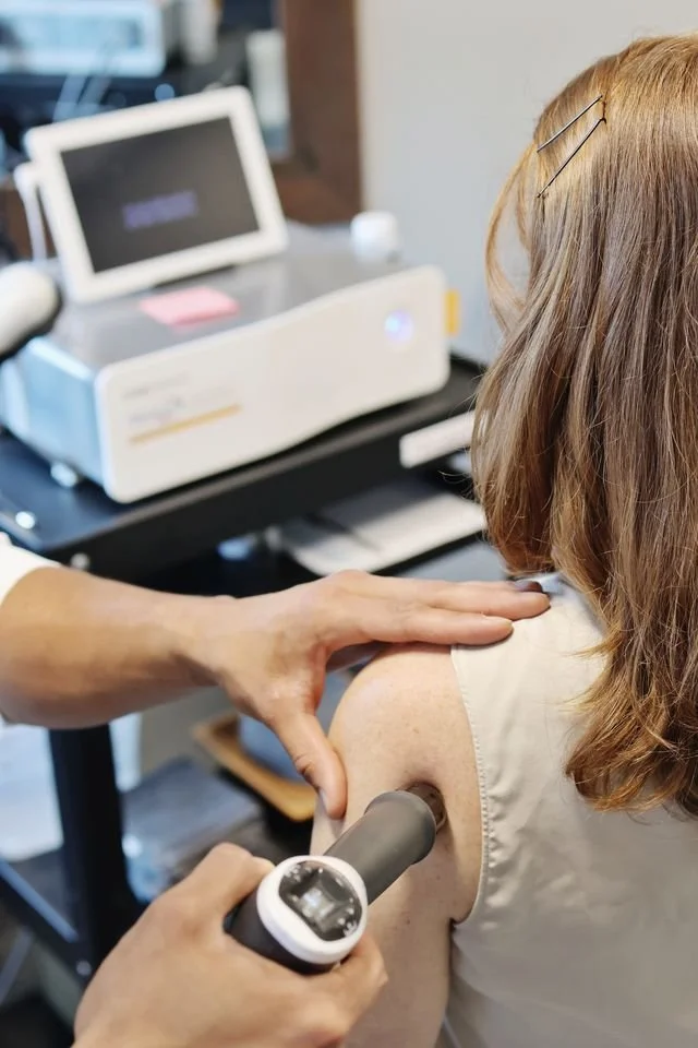 Patient receiving shockwave therapy in a chiropractic office, with a chiropractor giving the therapy  on the shoulder while a monitor and shockwave equipment are visible in the background.