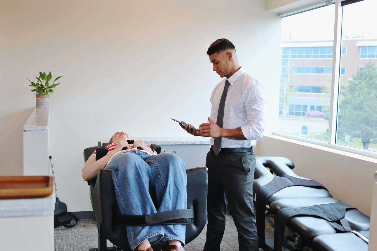 A young woman sitting in a chiropractic decompression chair with her legs supported by straps, relaxed with her eyes closed, while a male chiropractor hold the therapy device to begin treatment.