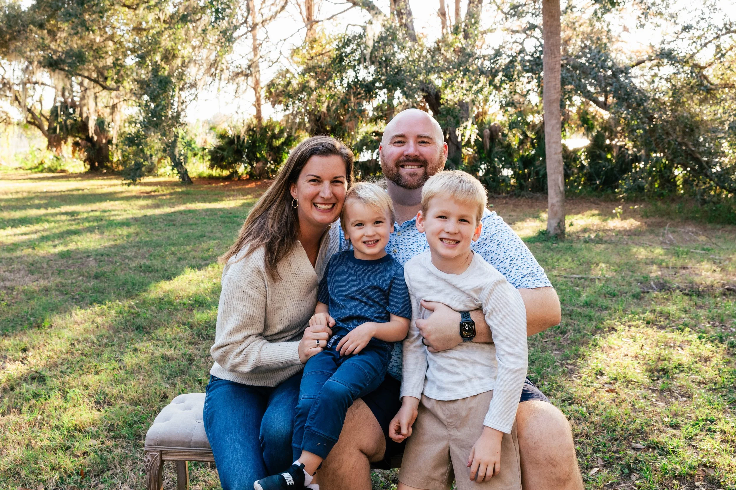 Family of four sitting on a bench in a grassy field during sunset, smiling.