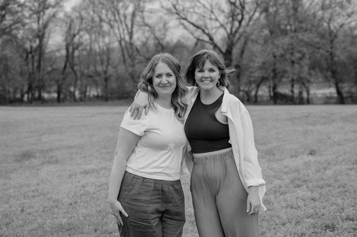 Two women standing side by side, one arm around the other's shoulder, smiling in a park with trees in the background, black and white photo.