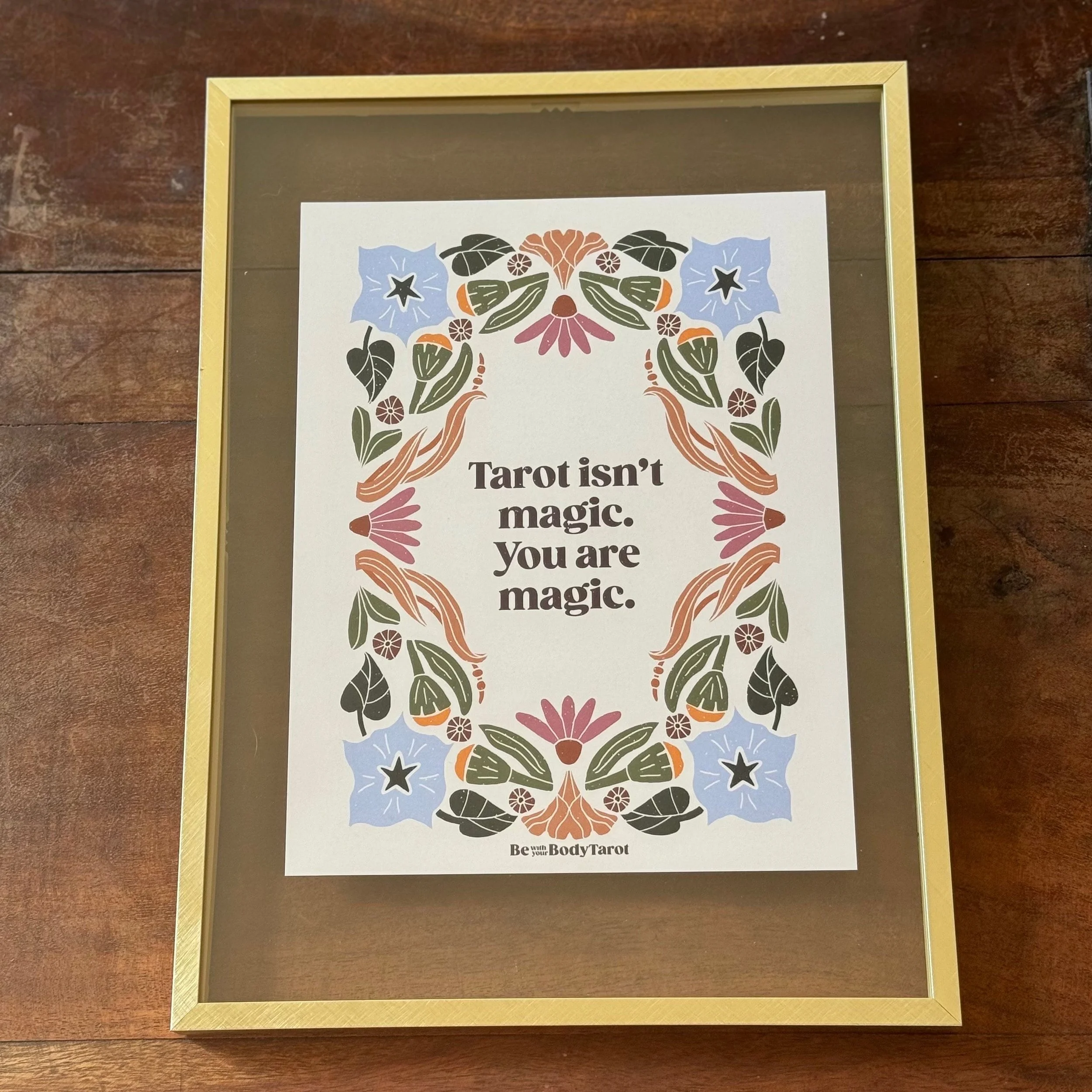 A framed print with a decorative border of floral and leaf illustrations surrounding the text, which reads 'Tarot isn't magic. You are magic.' at the center, with smaller text at the bottom saying 'Be your BodyTarot'.