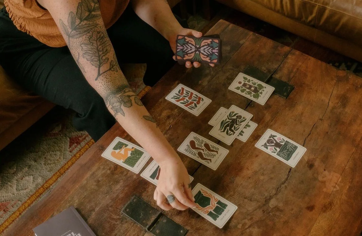 A person with tattoos on their arm holds Be With Your Body Tarot cards on a wooden table. Several cards with abstract nature-themed illustrations are spread on the table. A book or box is partially visible in the bottom left corner.