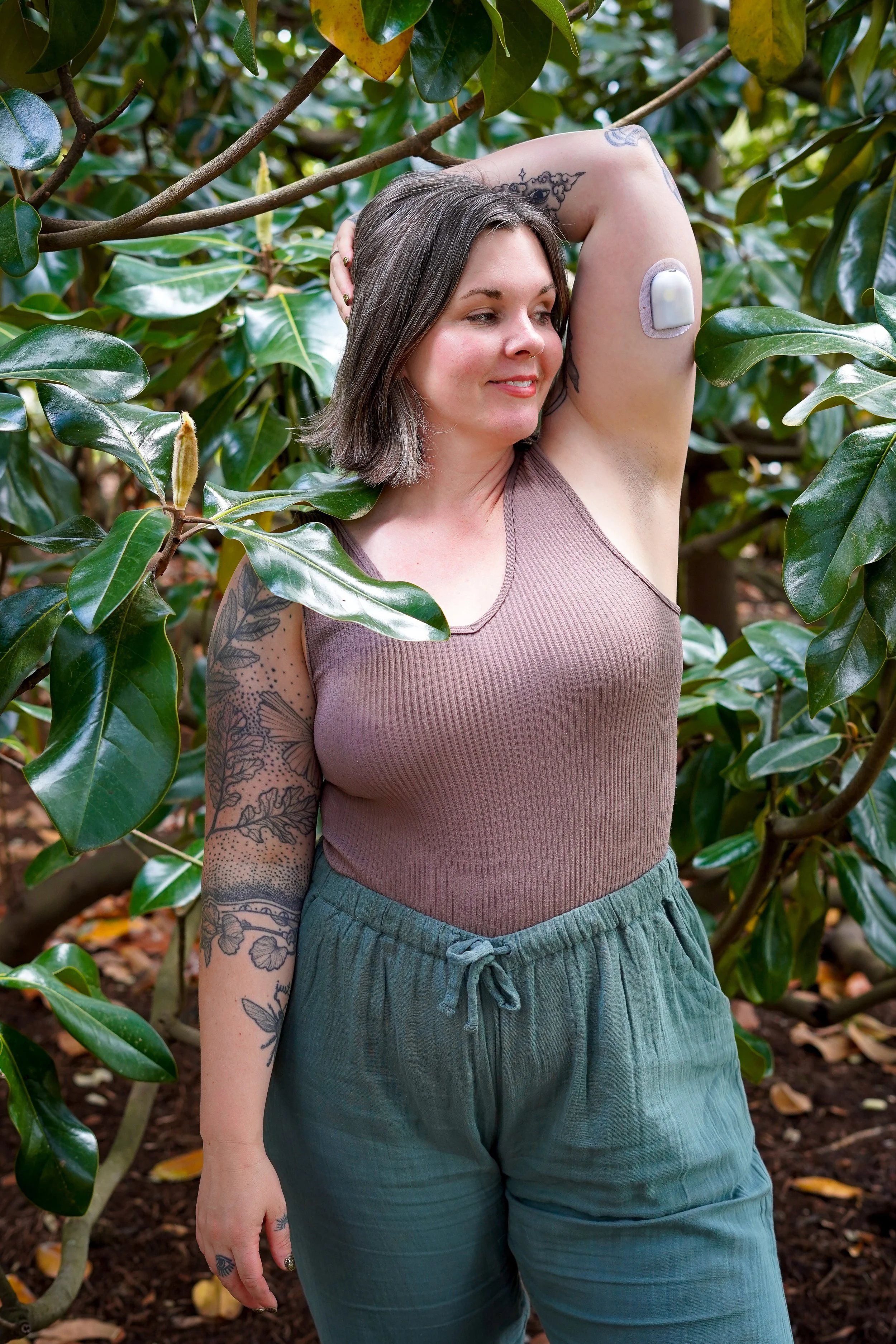 Sarah Jane standing in a garden, wearing a sleeveless top with tattoos on their arm, and displaying a medical device on their upper arm.