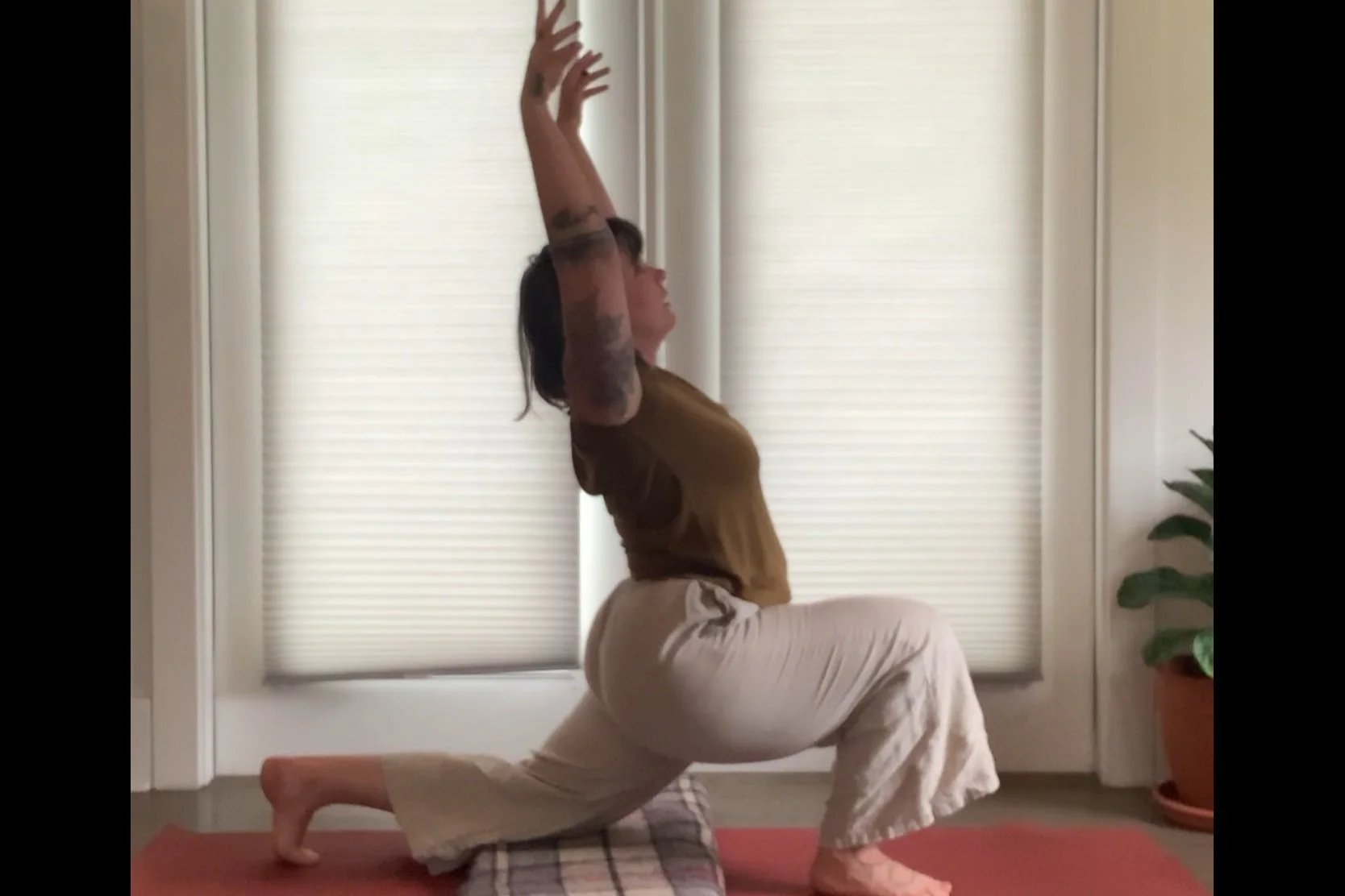 Person practicing yoga indoors in a low lunge pose on a red mat beside a potted plant.