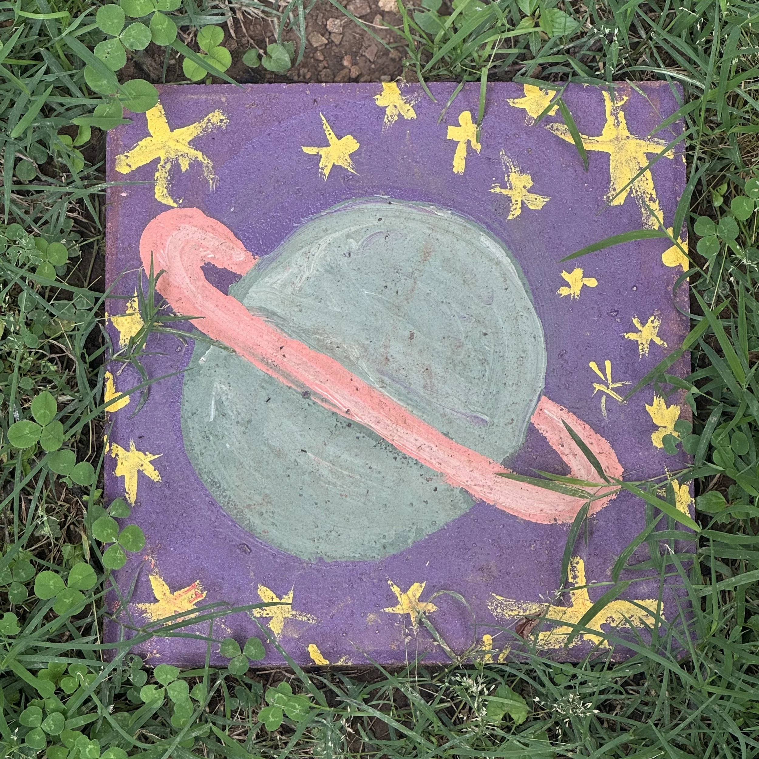 A painted square tile with a cosmic theme, featuring a green planet with a pink ring, yellow stars, and a purple background, surrounded by grass and clover leaves.