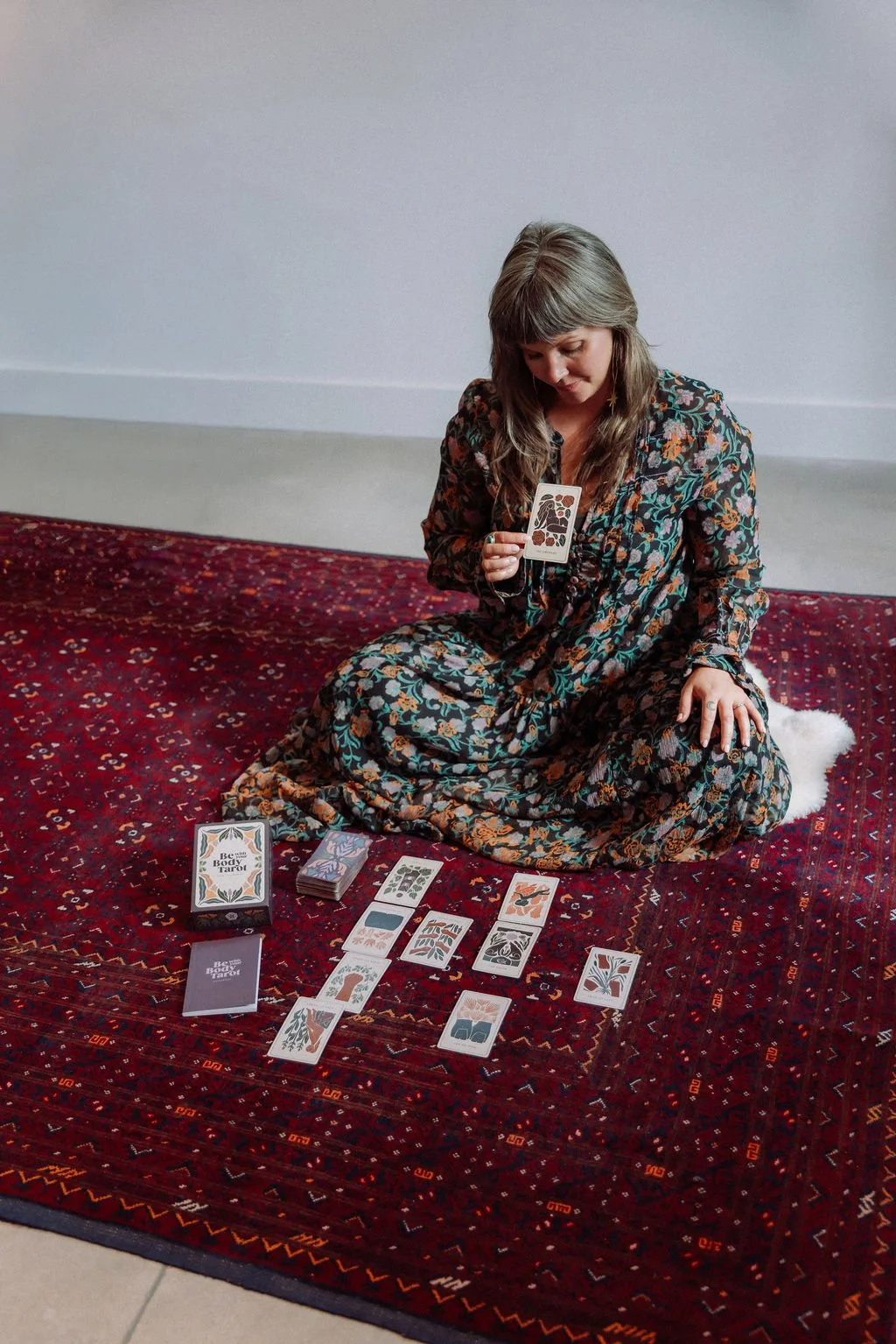 Sarah Jane seated on a red rug holding a tarot card above a tarot spread