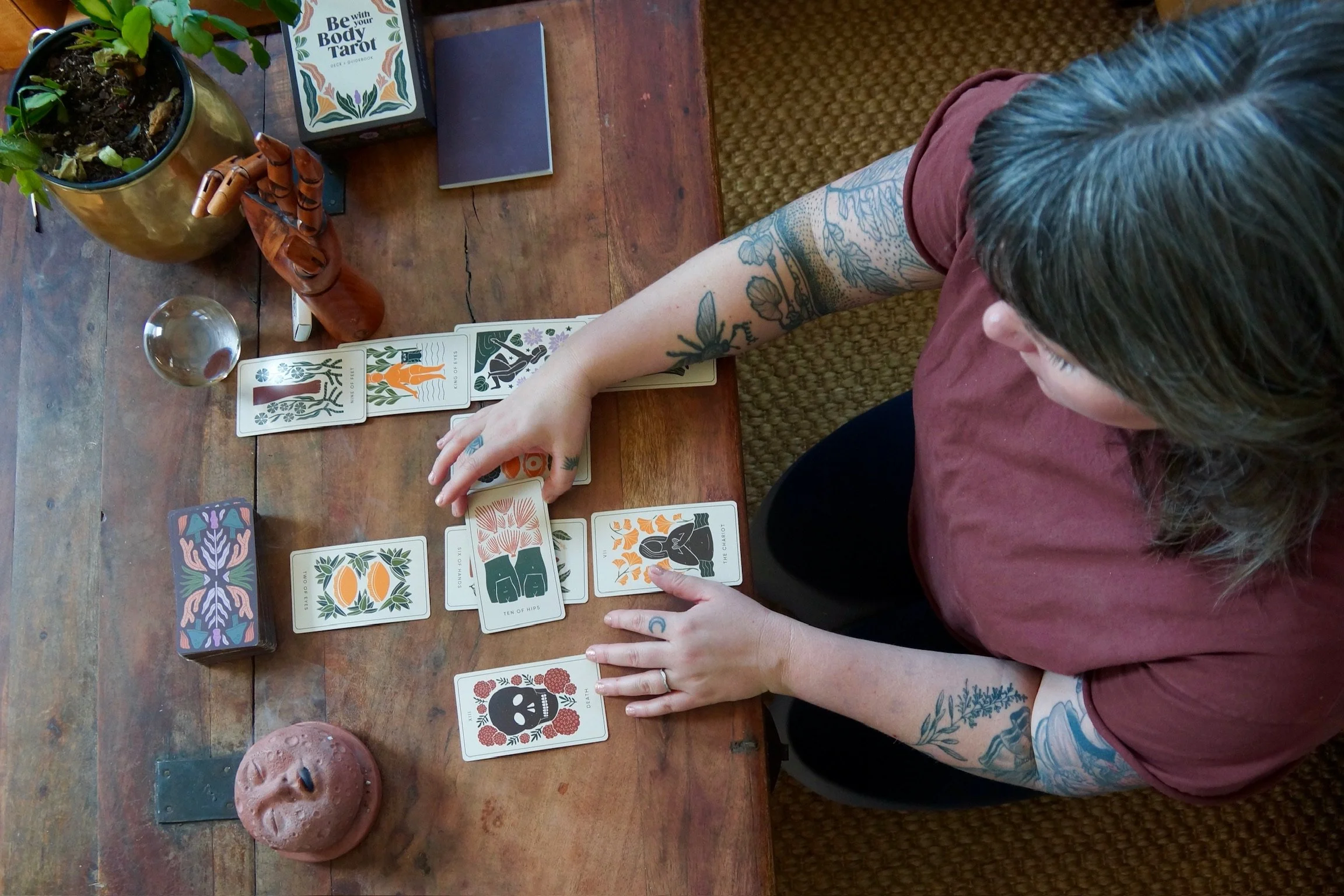 A person with tattoos and long hair is sitting at a wooden table playing a tarot card game. The table has plants, a crystal ball, a small skull-shaped object, and tarot cards with colorful illustrations on it.