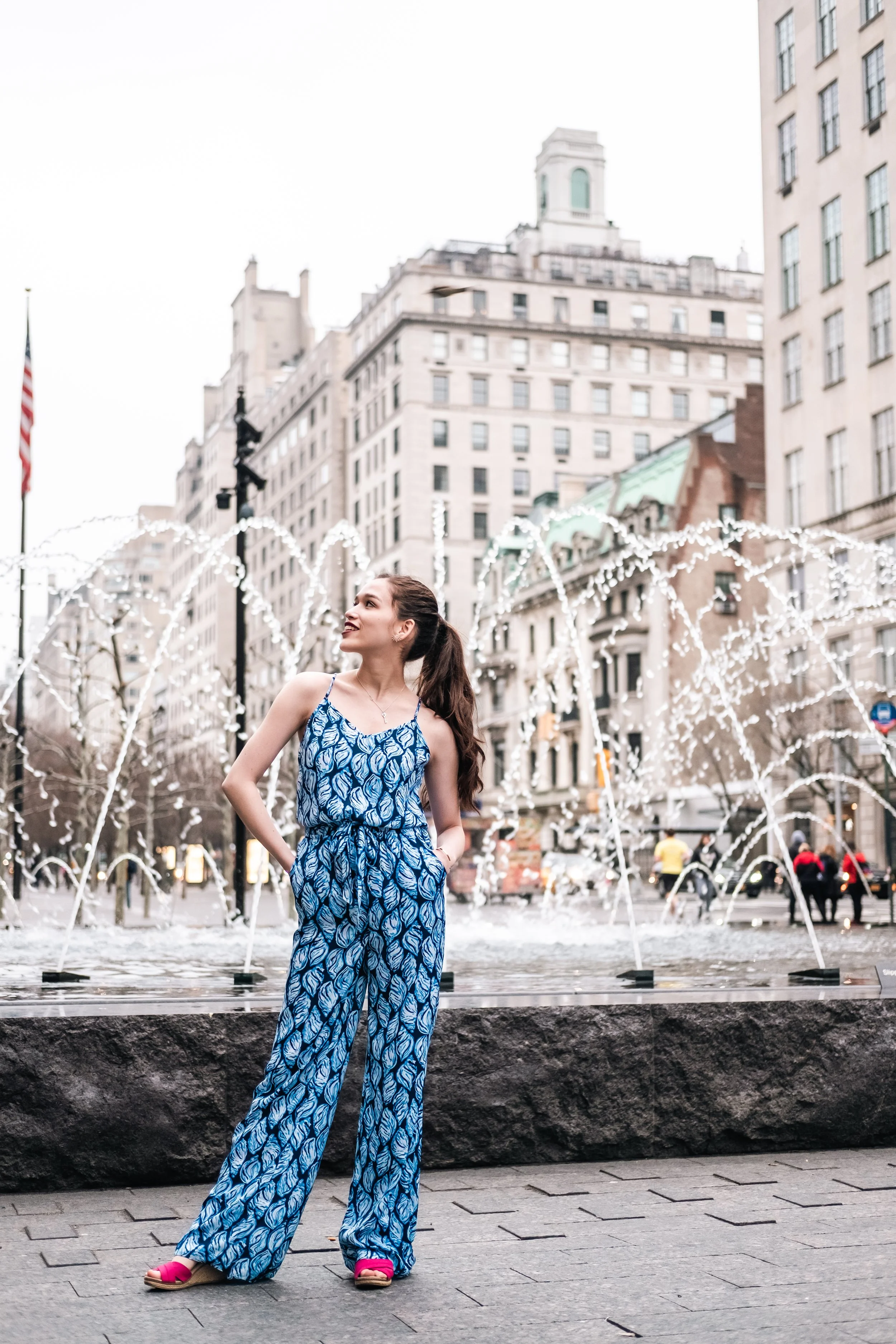 A woman in a blue patterned jumpsuit and pink sandals posing in front of a fountain and city buildings.
