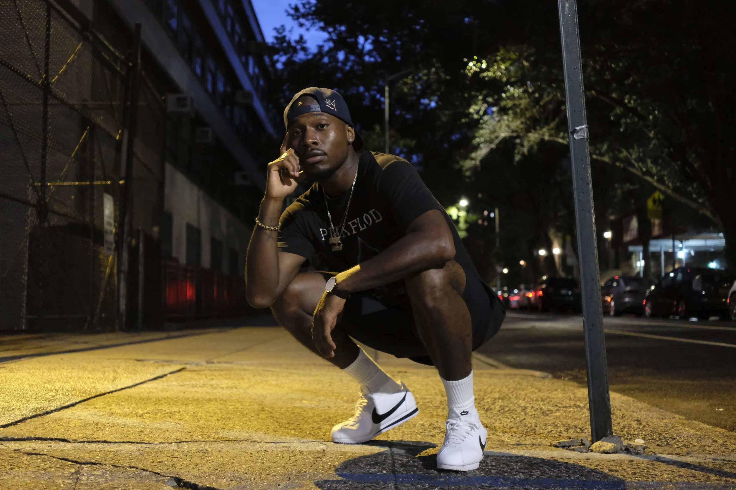 A young man wearing a backward cap, a black t-shirt, white Nike sneakers, and jewelry is squatting on a city sidewalk at night, resting his chin on his hand, with cars and trees in the background.