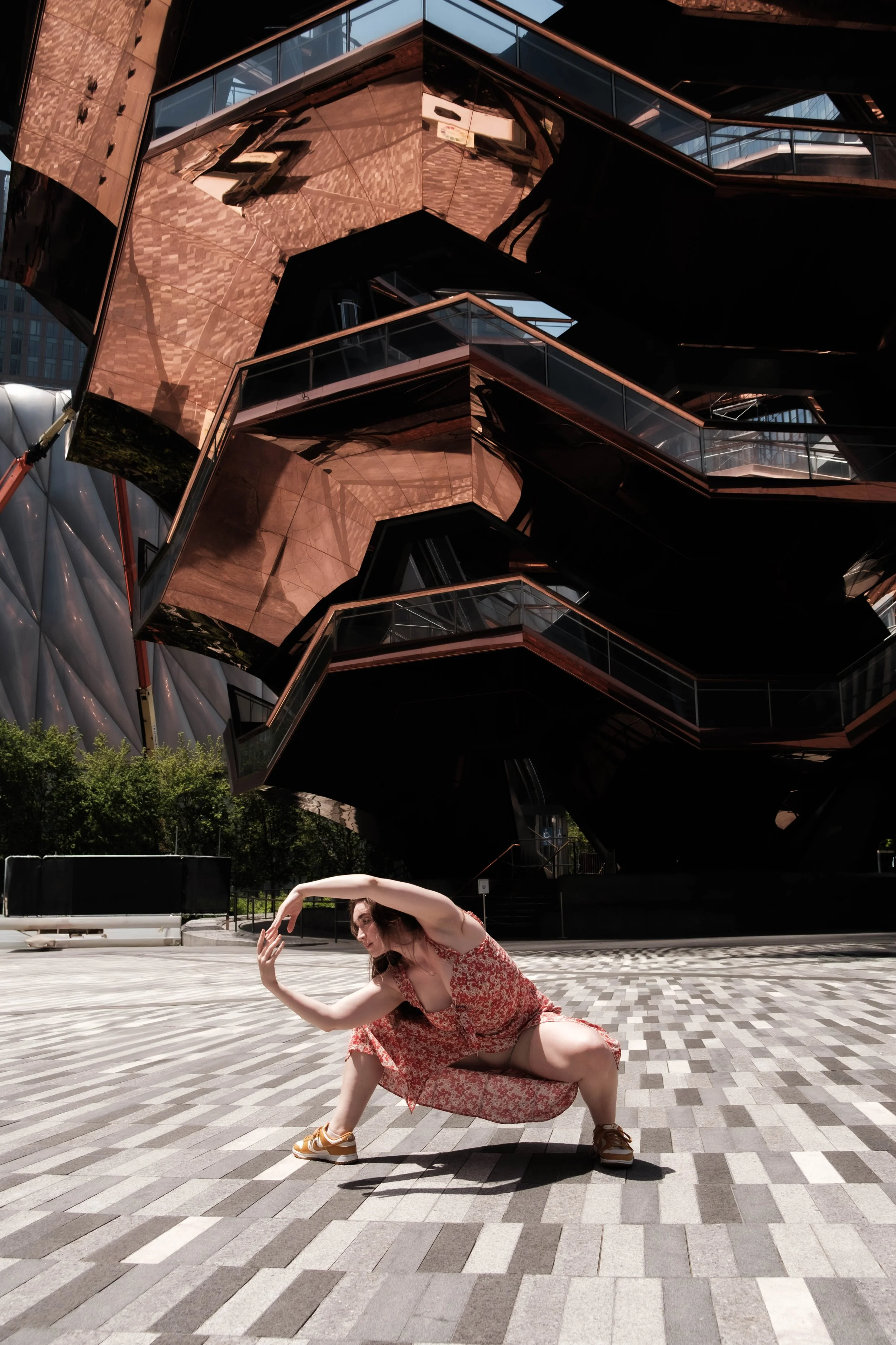 A woman in a floral red dress and yellow sneakers is performing a dance pose on a patterned outdoor plaza surface, with modern building architecture featuring reflective surfaces and glass railings in the background.
