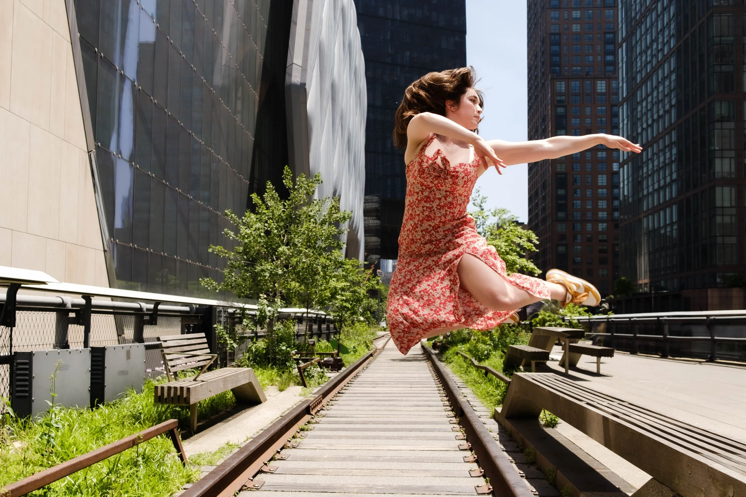A woman in a red floral dress is jumping between train tracks in an urban park, with skyscrapers in the background.
