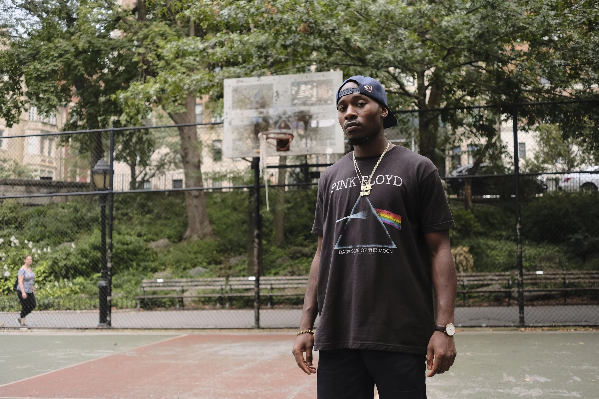 A young man with dark skin wearing a dark Pink Floyd t-shirt, gold chain, a wristwatch, and a backward navy baseball cap stands on an outdoor basketball court with a basketball hoop and tall trees in the background. People are walking nearby.