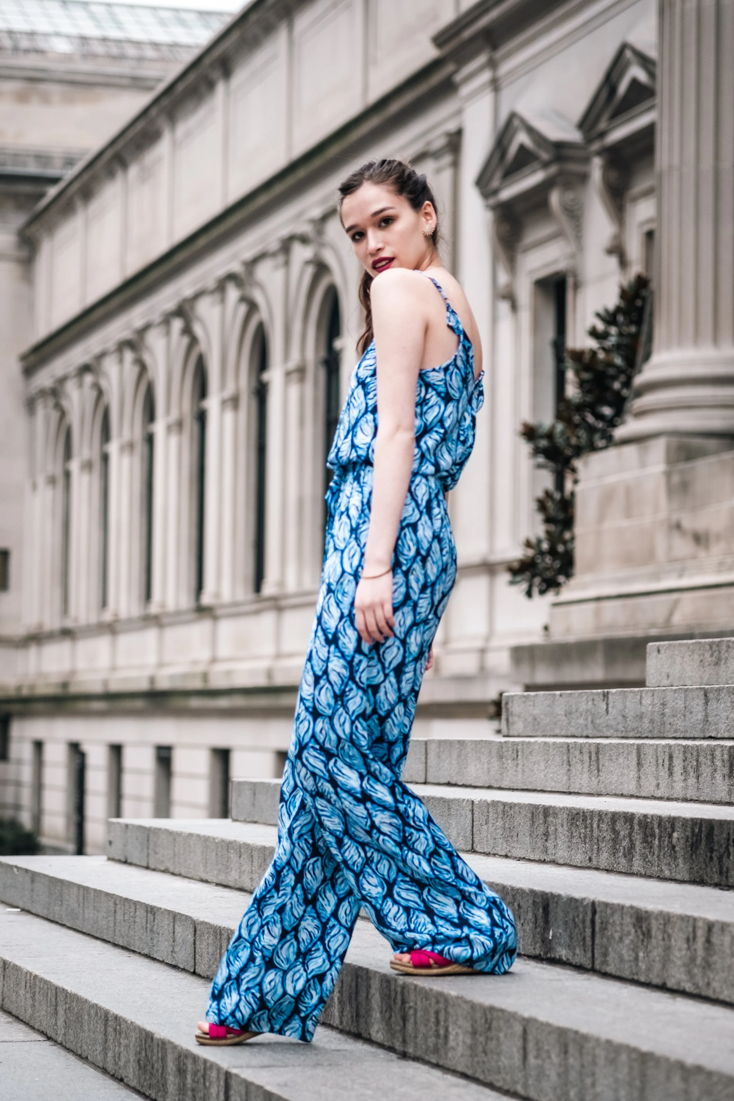Young woman in a blue patterned jumpsuit and pink sandals walking down outdoor stone steps in front of a classical building with arched windows and decorative columns.