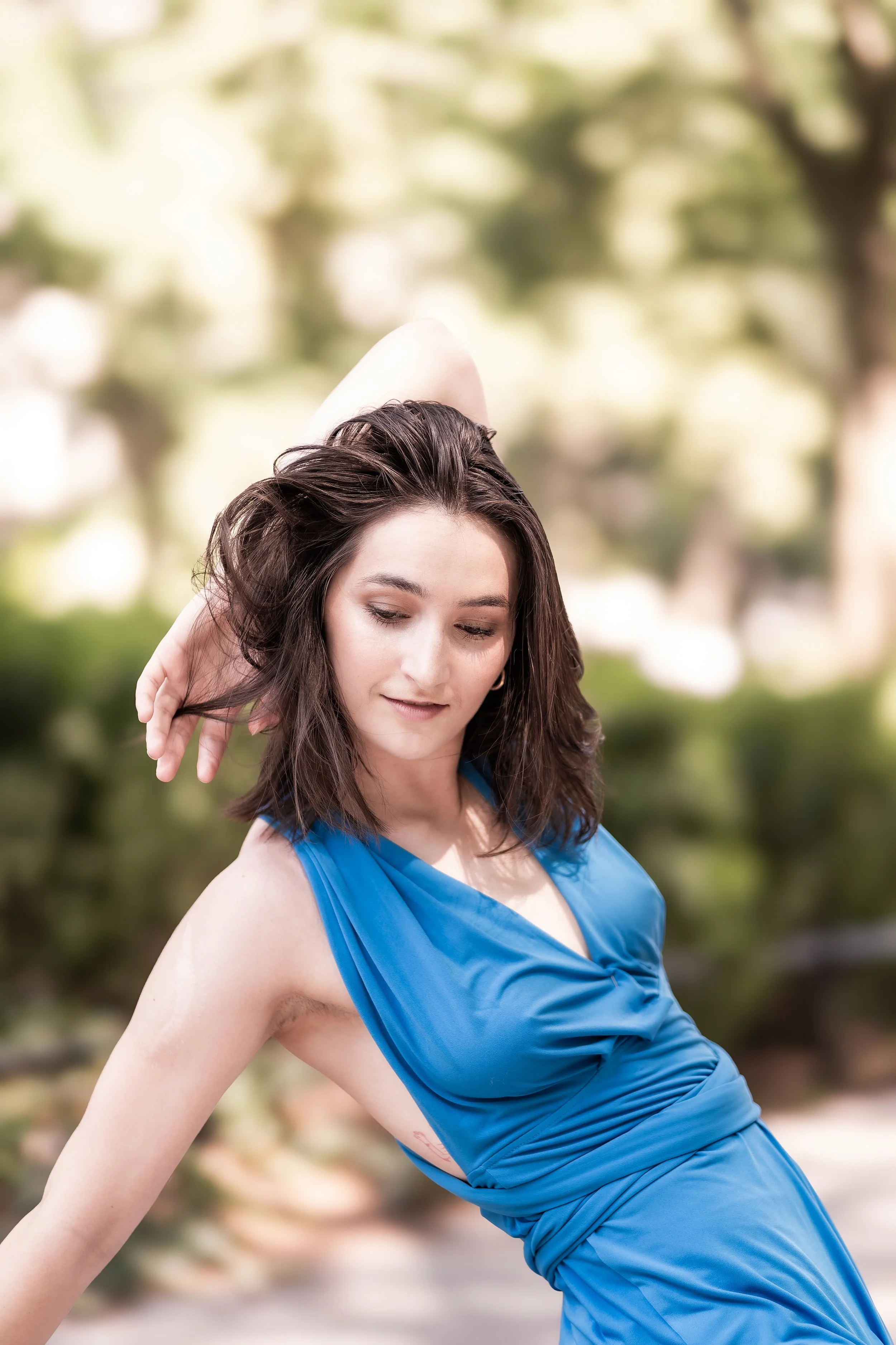 A woman in a blue dress practicing yoga outdoors with her eyes closed and a serene expression, surrounded by a blurred green and tan background of trees.