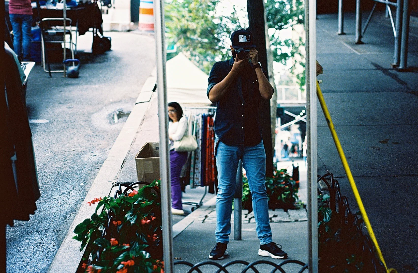 Person taking a photo of themselves in a mirror at an outdoor market, with a woman shopping and various market stalls in the background.