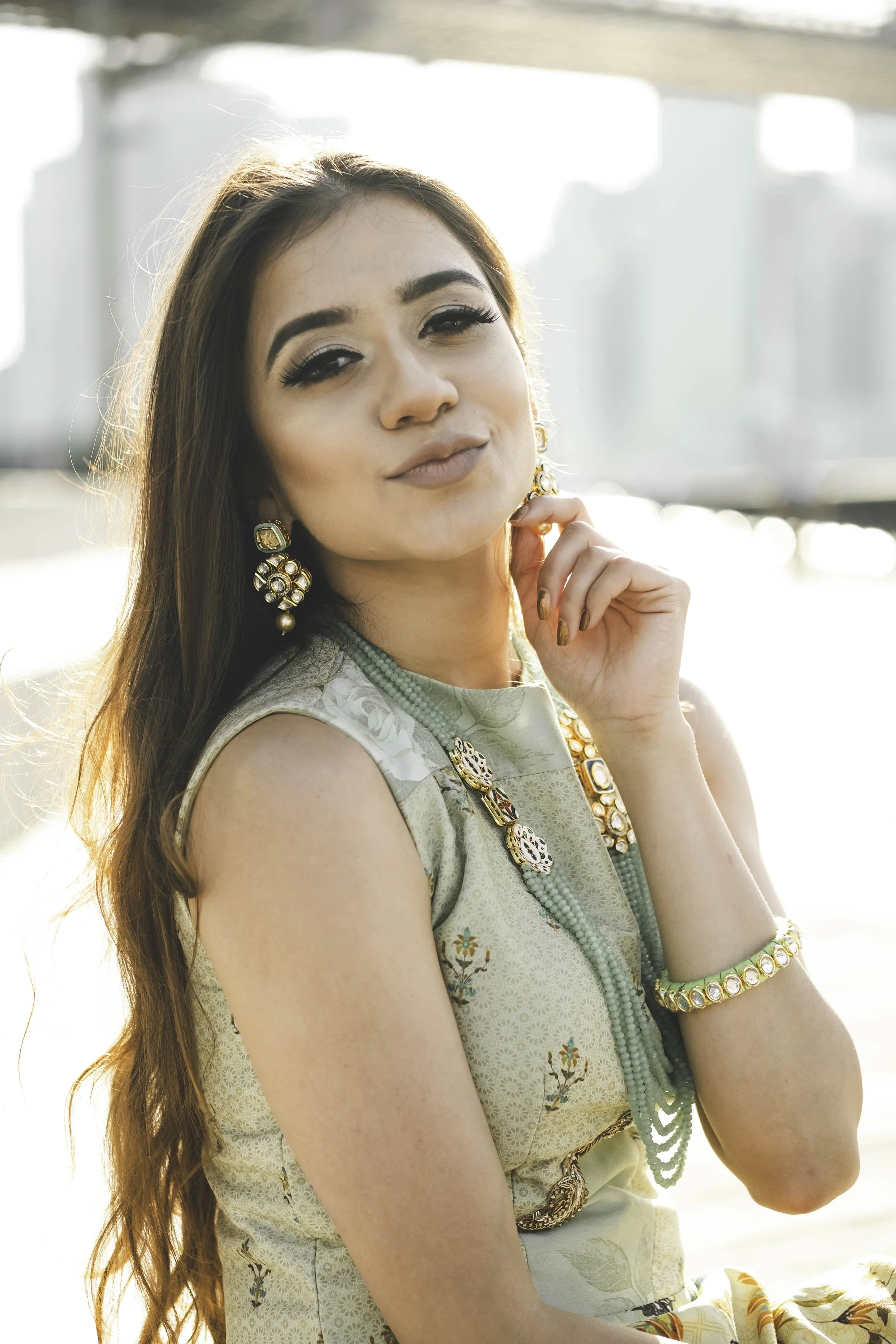 A woman with long brown hair, wearing makeup, jewelry, and a traditional dress, is smiling slightly and holding her hand near her chin outdoors during daylight with a blurred background.
