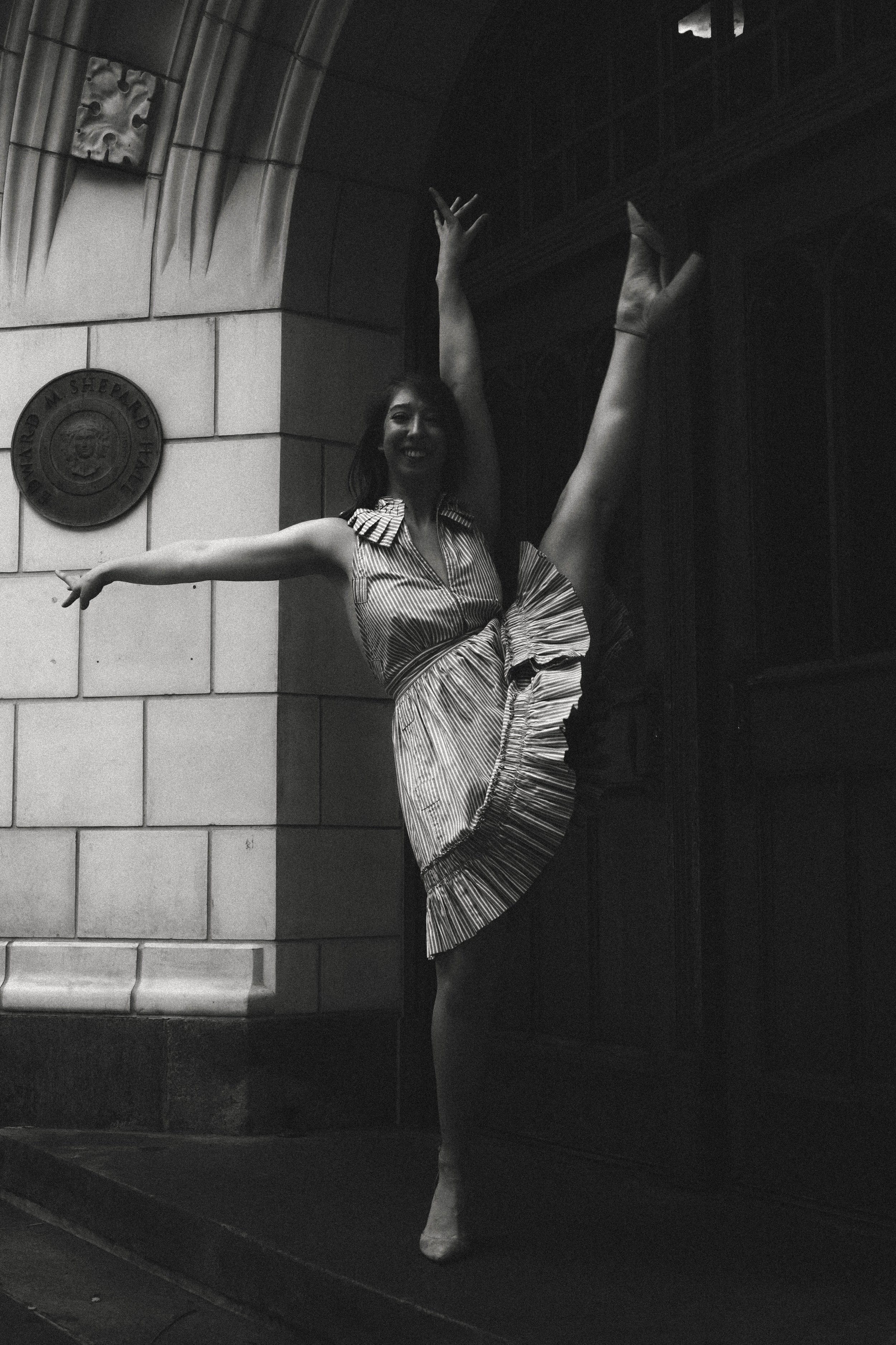 A woman in a striped dress is performing a high leg ballet pose on stage with one arm raised and one extended to the side, against a backdrop of a brick wall with a circular emblem and an archway.