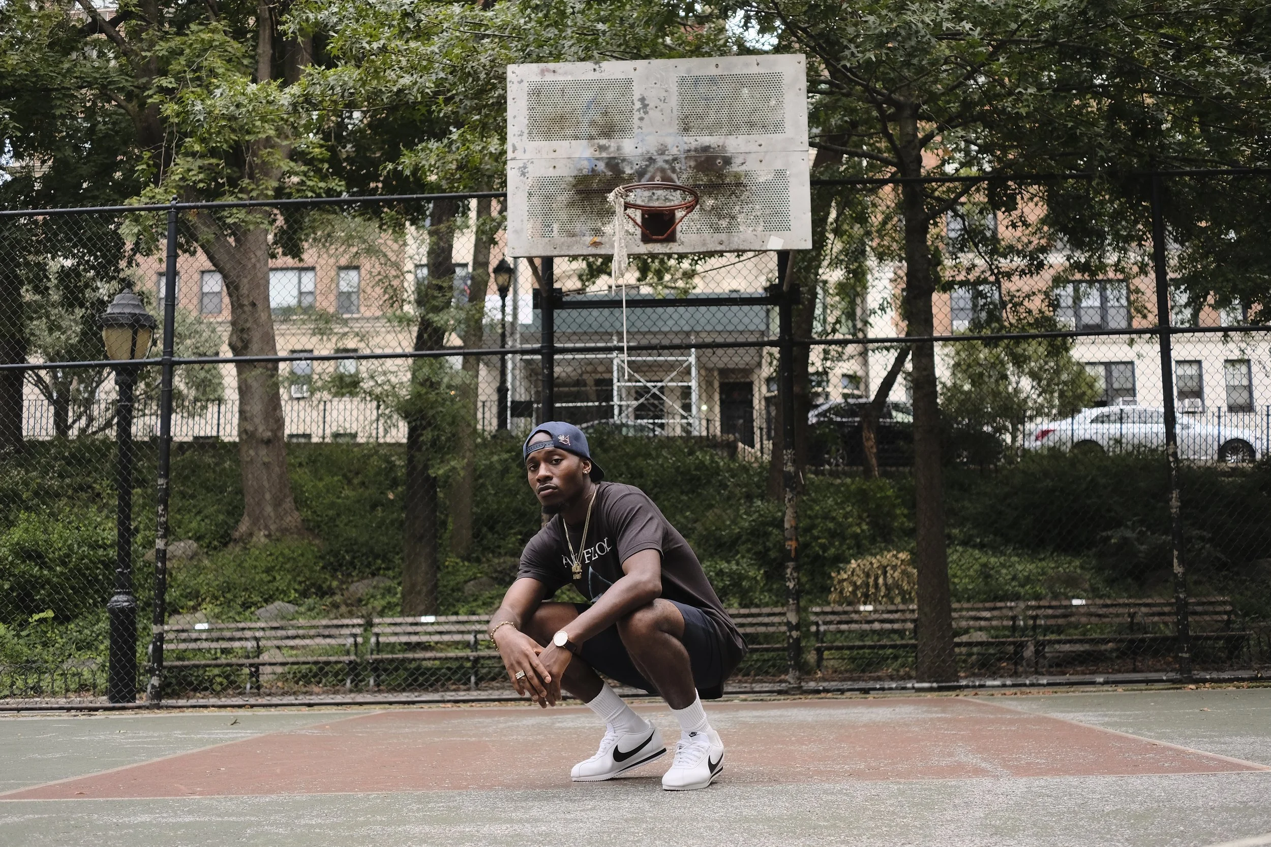 Young man in athletic clothing crouching on a basketball court, with a weathered backboard and hoop behind him, surrounded by trees and a chain-link fence.