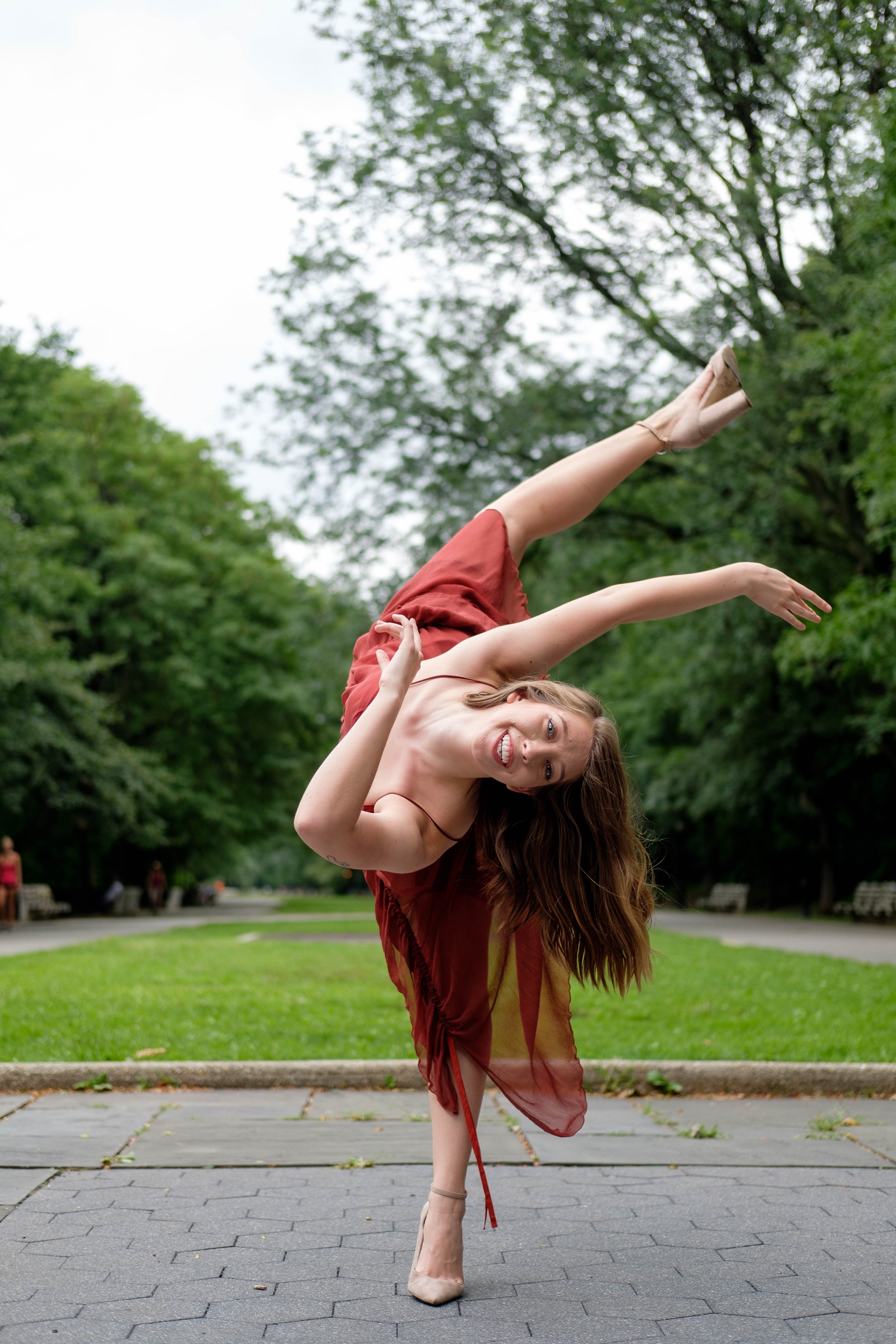 A woman with long brown hair dancing outdoors in a park, wearing a flowy red dress and beige high heels, with trees and benches in the background.
