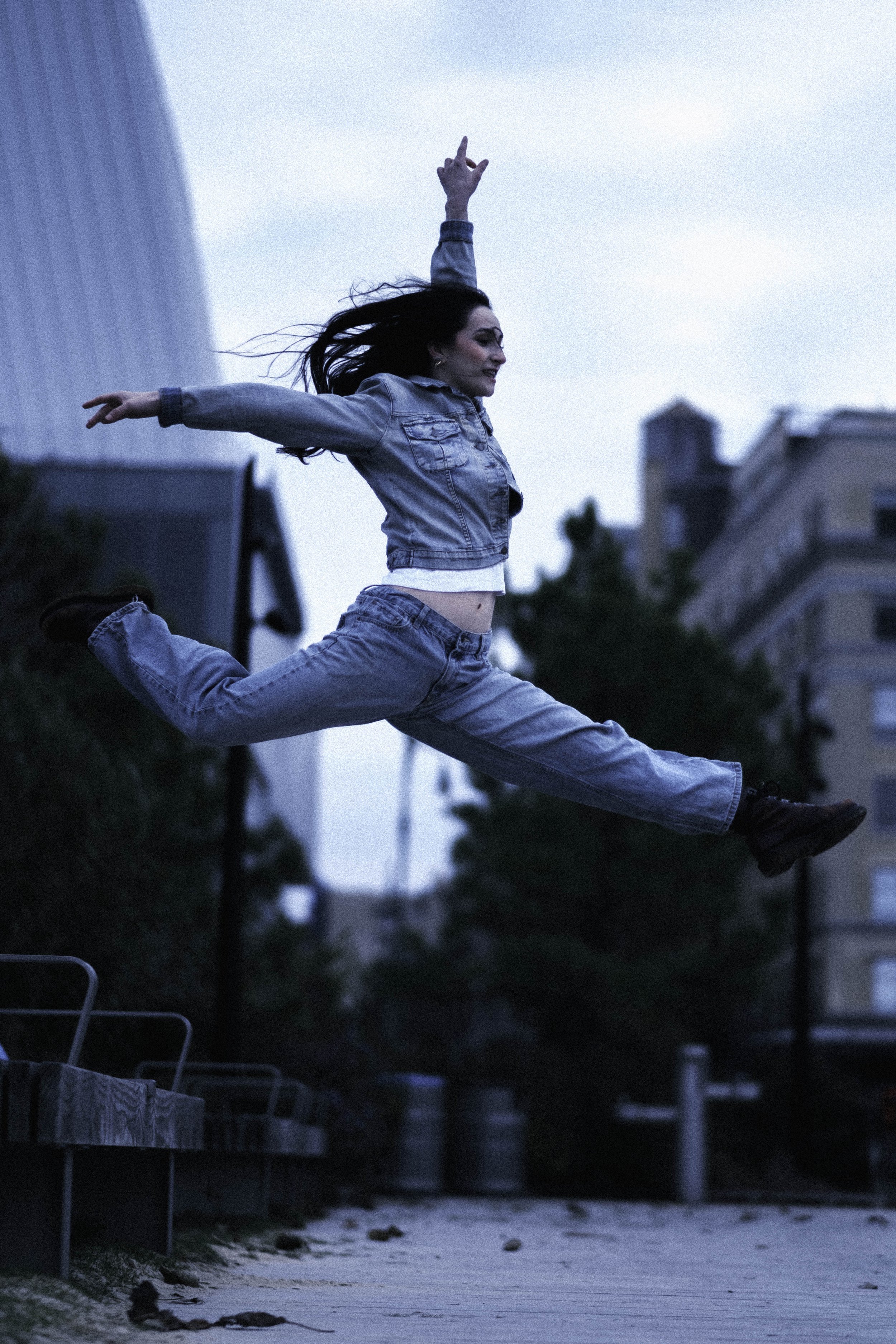 A woman in jeans and a denim jacket is mid-air in a jump, with her arms and legs extended outward, against an urban backdrop during dusk or early evening.