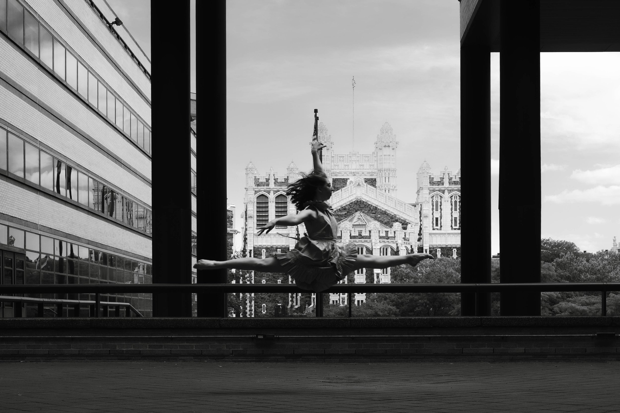 A ballet dancer performing an arabesque on an outdoor stage with a cityscape background, black and white photo.