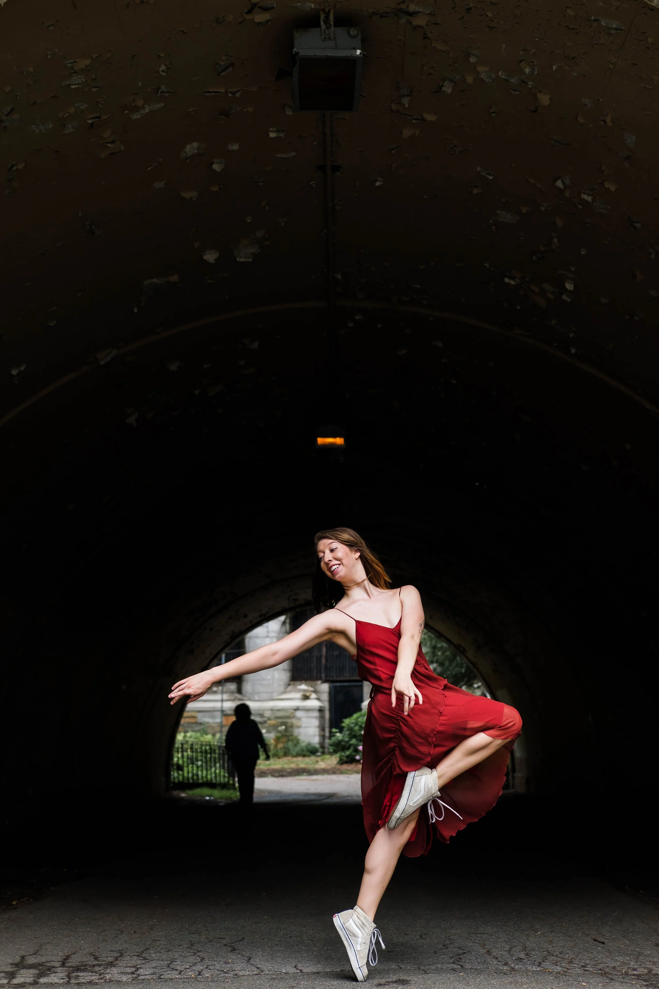 A woman in a red dress dancing in a tunnel with a person walking in the background.