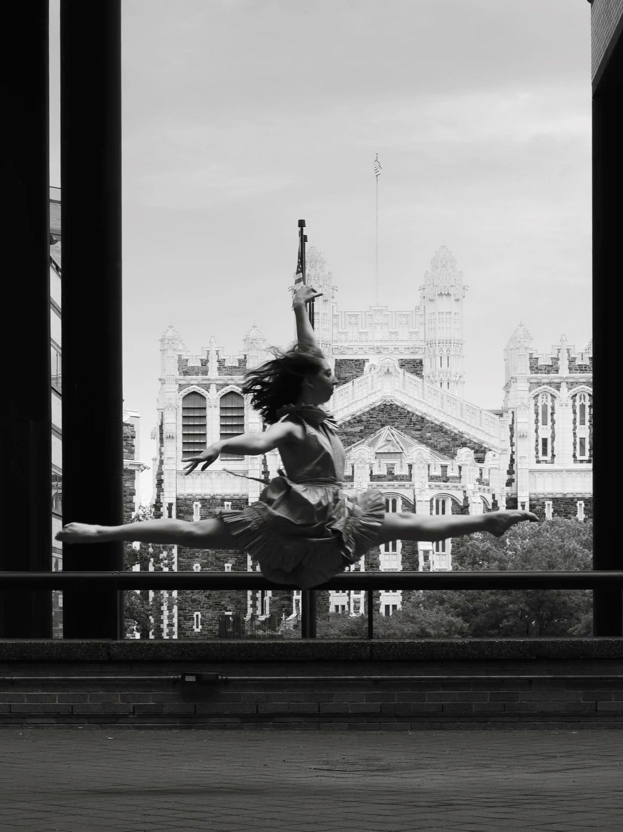 A ballet dancer performing a grand jeté on a stage with a historic building and flag in the background.