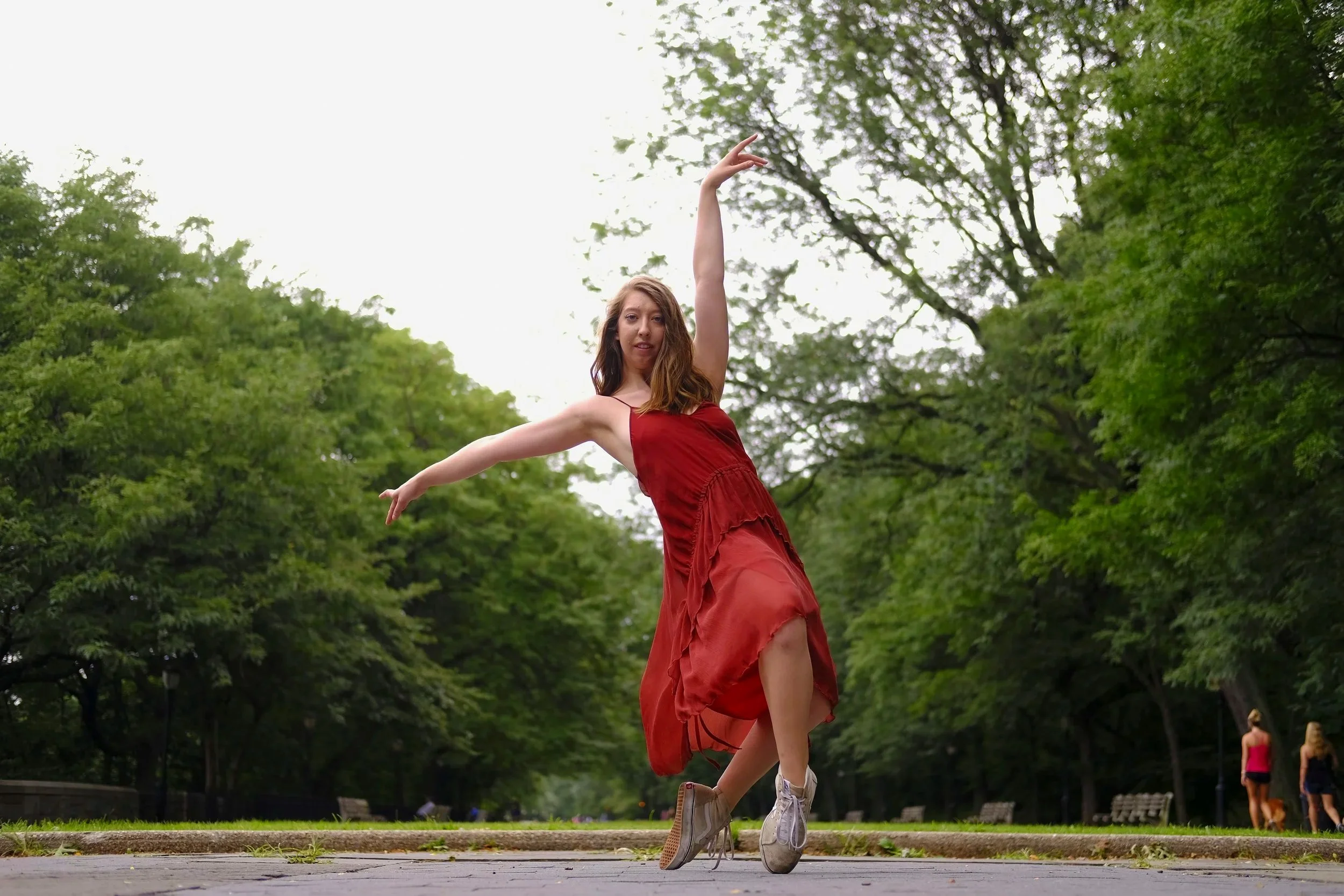 A woman in a red dress dancing outdoors on a park path surrounded by green trees.