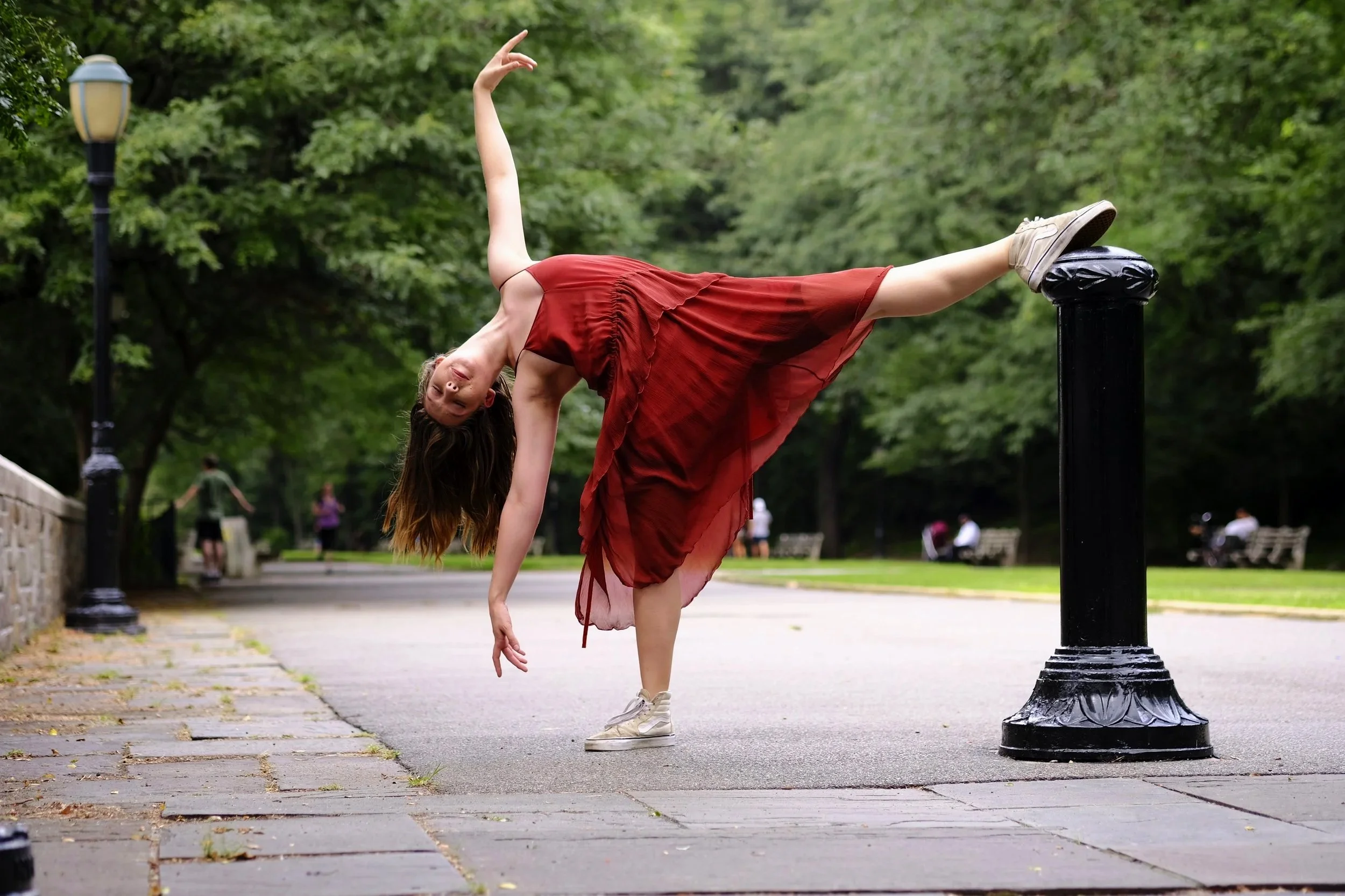 A woman in a red dress performing a handstand with one leg resting on a black post in a park with green trees and walking paths.