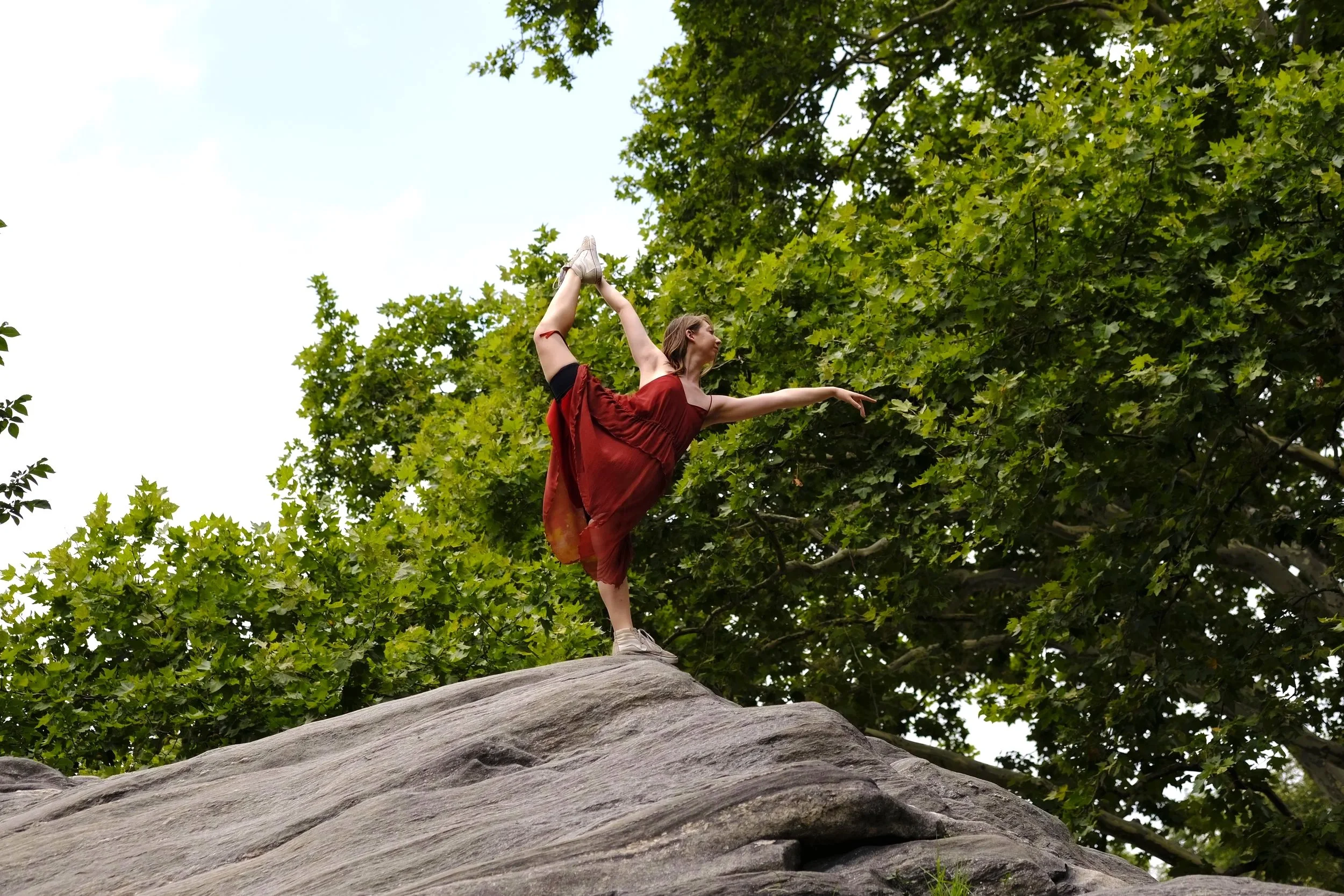 A woman in a red dress performing a yoga pose on a tree trunk in a park with green leaves.