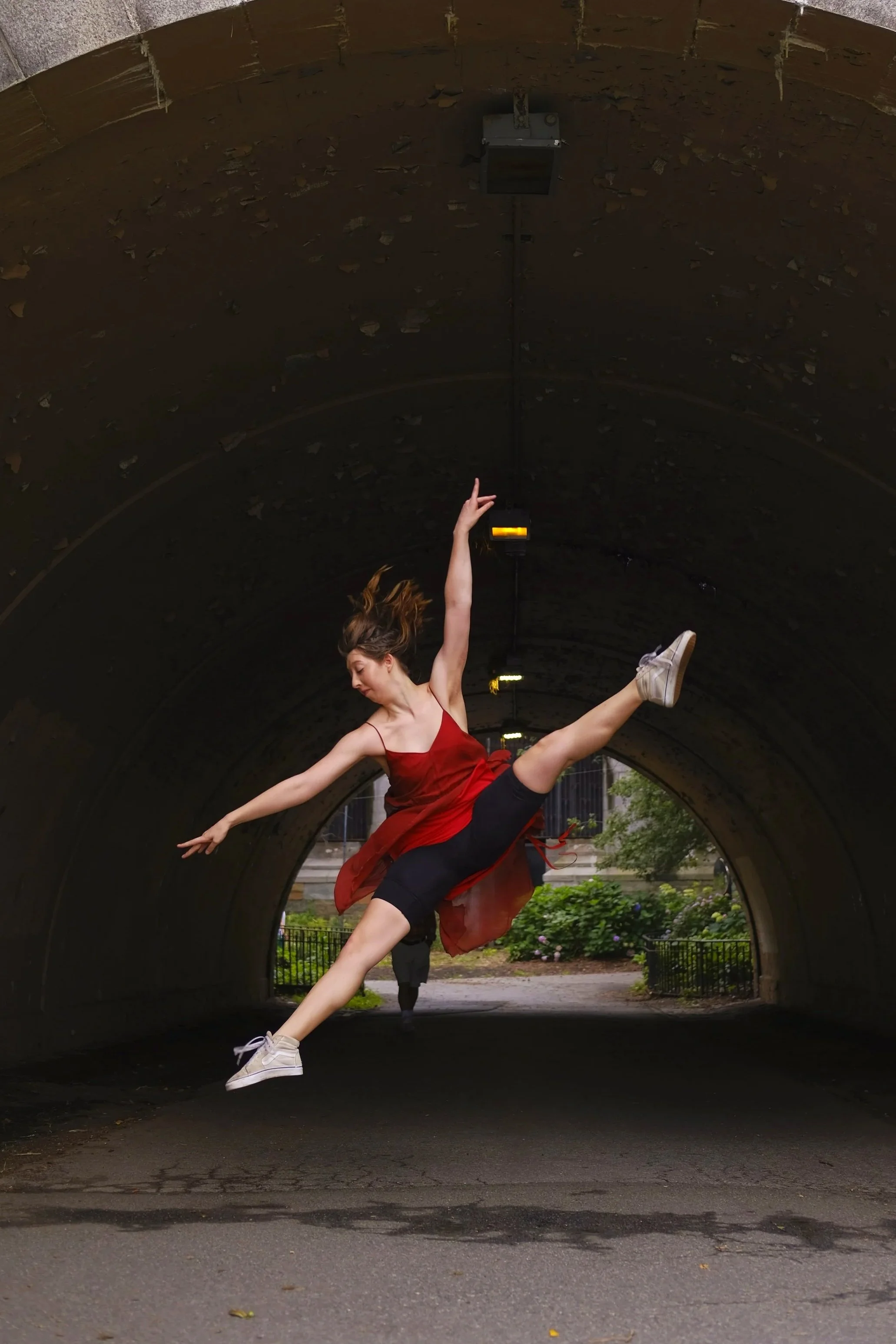 A dancer in a red dress and white sneakers is doing a leap inside a tunnel with a dark, textured interior. There are streetlights and greenery visible outside the tunnel.