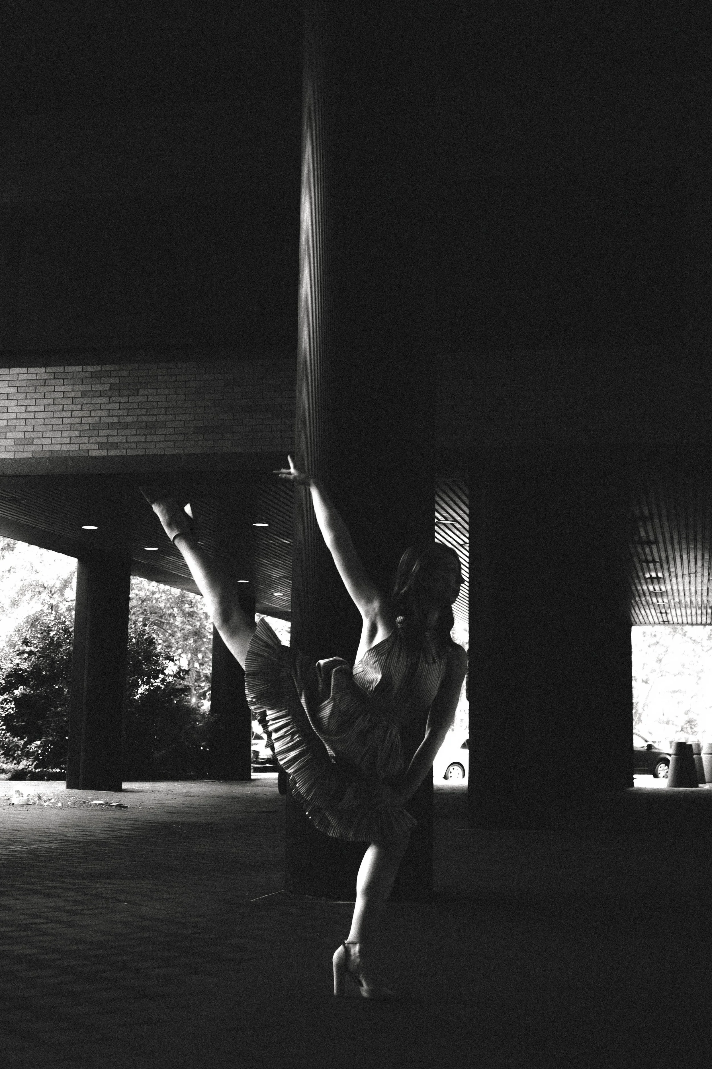 A woman in a ballet pose, balancing on one leg and holding her other leg up with her arm, wearing a striped dress and pointe shoes, under a covered outdoor area with support pillars and a brick building in the background.