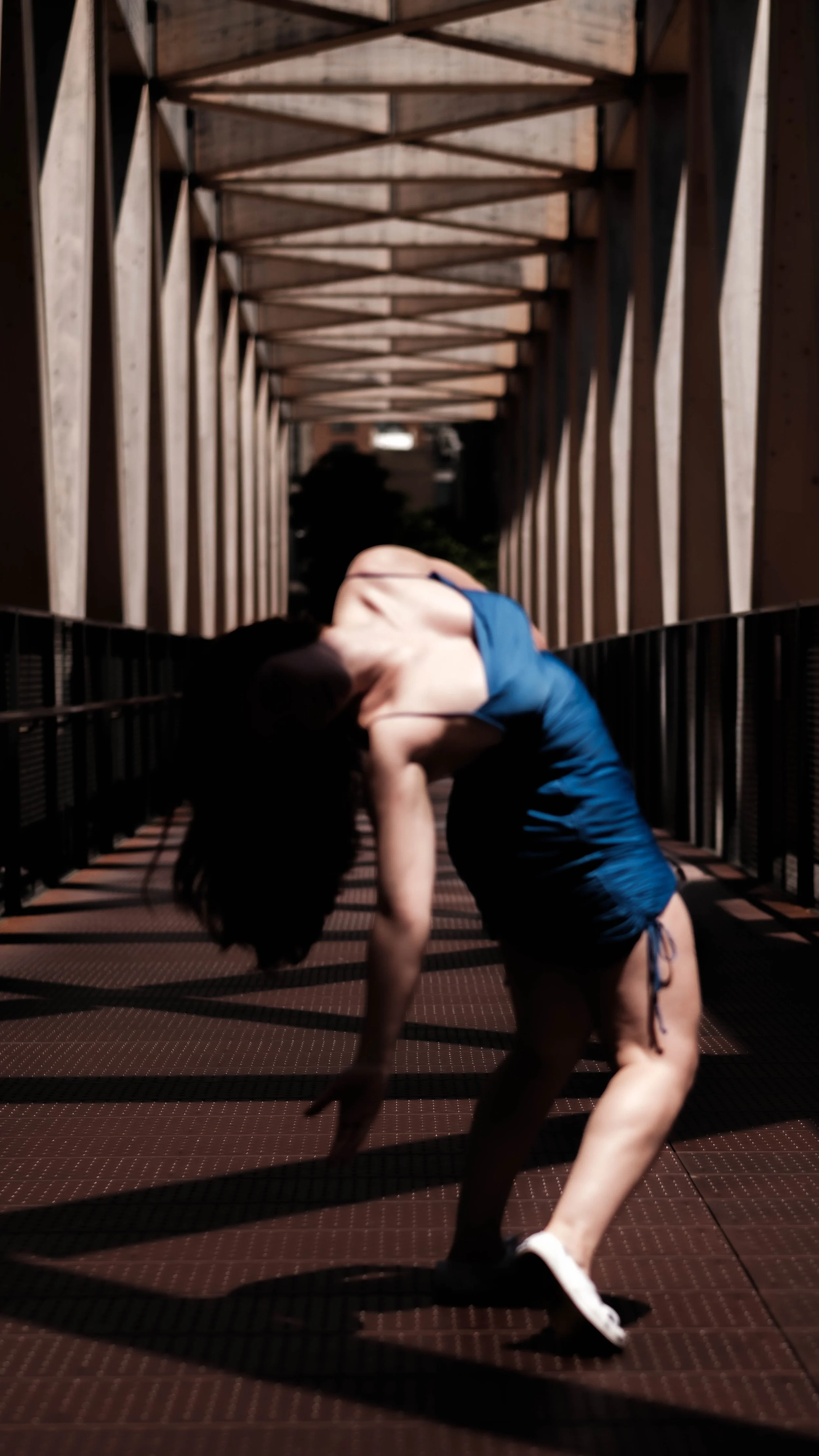 A woman in a blue dress is performing a dance or yoga pose on an outdoor walkway with a wooden geometric bridge structure overhead at night.