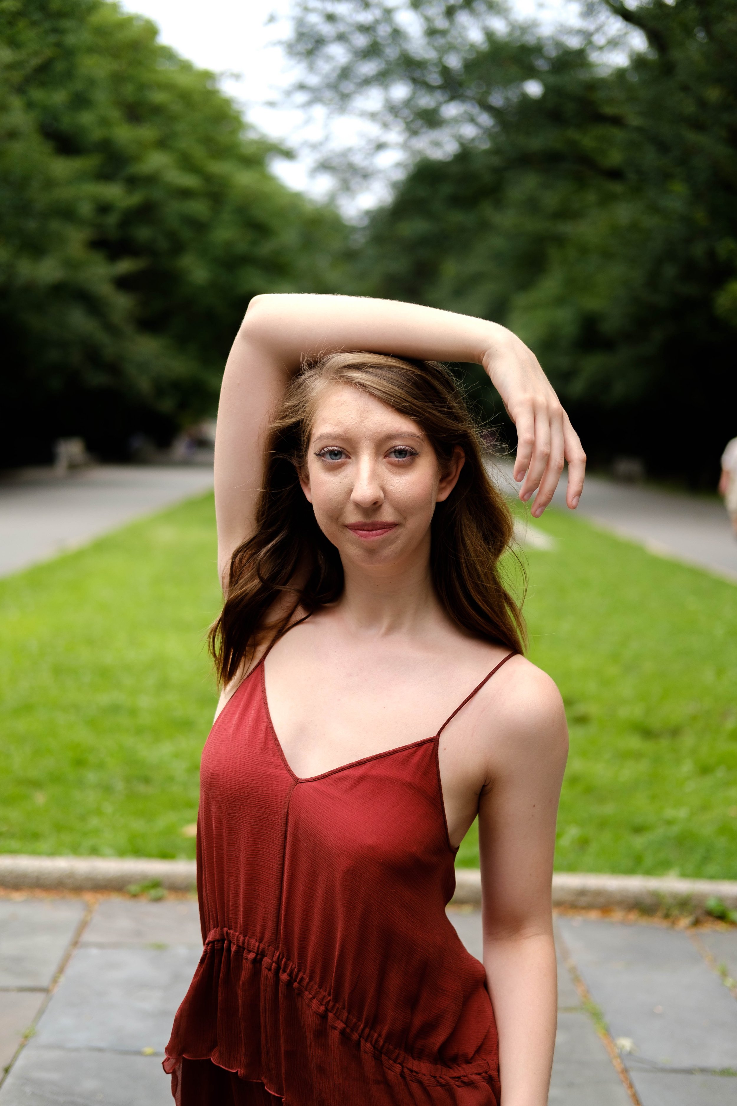 Young woman with fair skin and long brown hair standing outdoors on a sidewalk in front of green trees, wearing a sleeveless red dress with spaghetti straps.