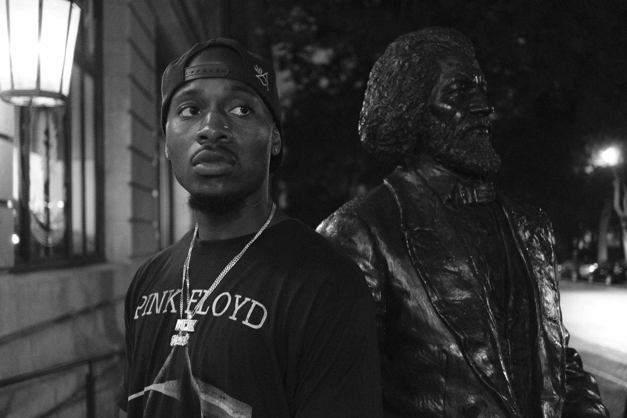 A young man wearing a black baseball cap backward and a Blackpink Floyd t-shirt stands next to a bronze bust of a man with distinctive hair and facial features at night.