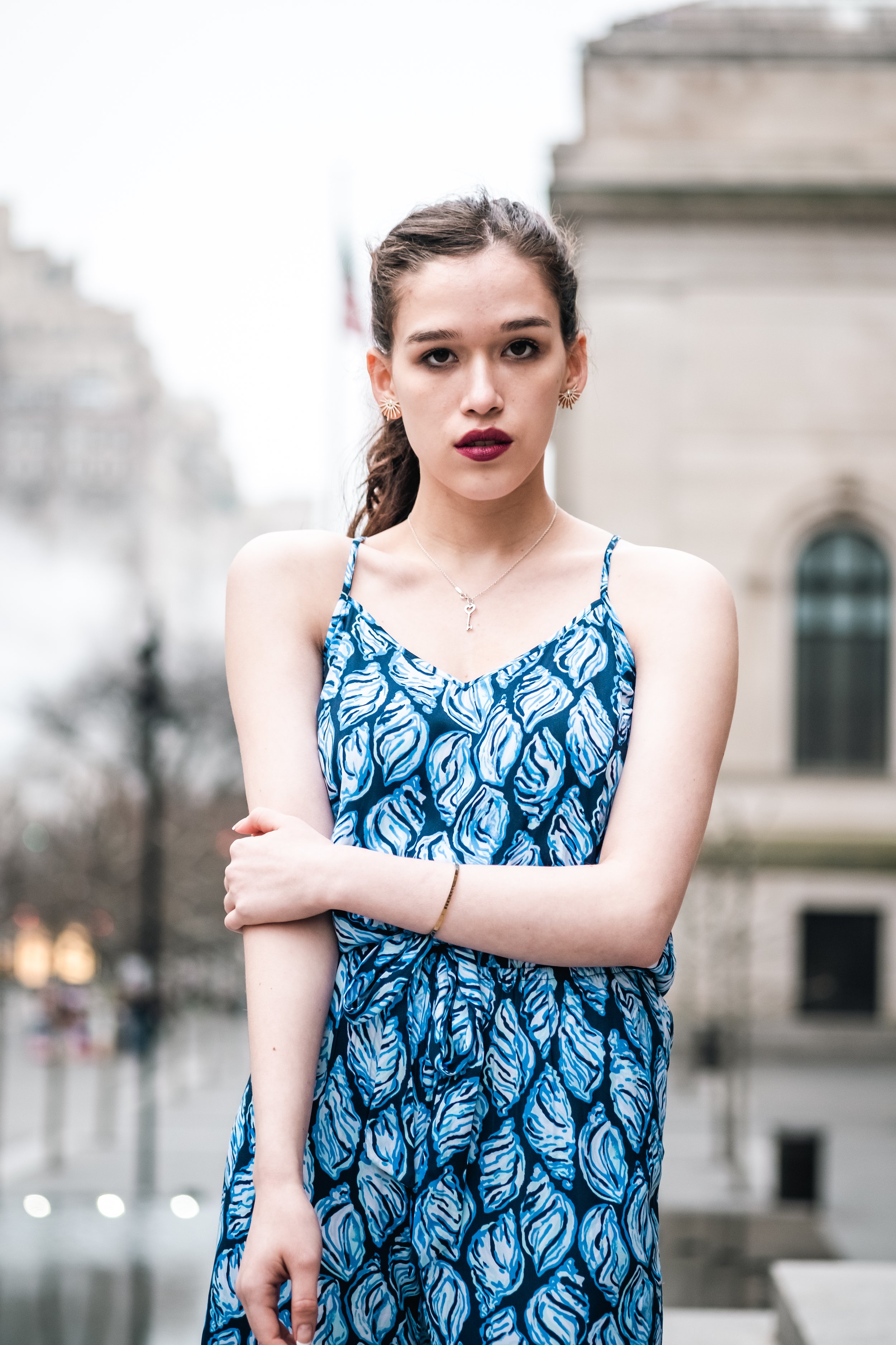 A young woman with brown hair in a ponytail, wearing a blue and white patterned sleeveless dress, standing outdoors with buildings and a blurred city background, crossing her arms.