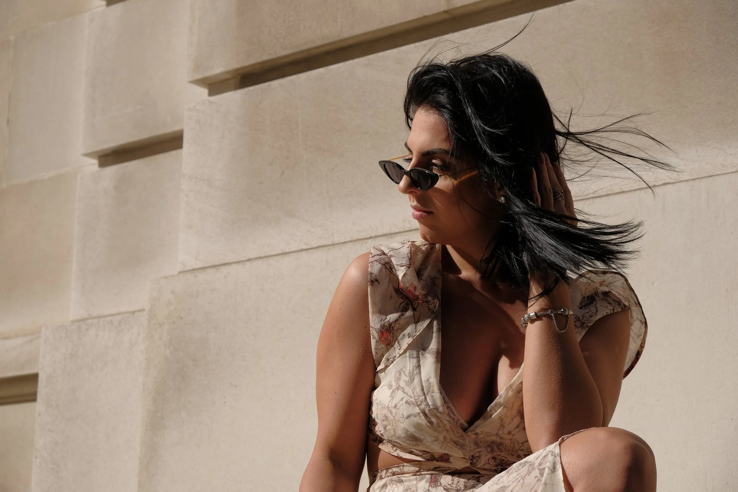 A woman with dark hair and sunglasses sitting against a beige wall, wearing a floral sleeveless dress.