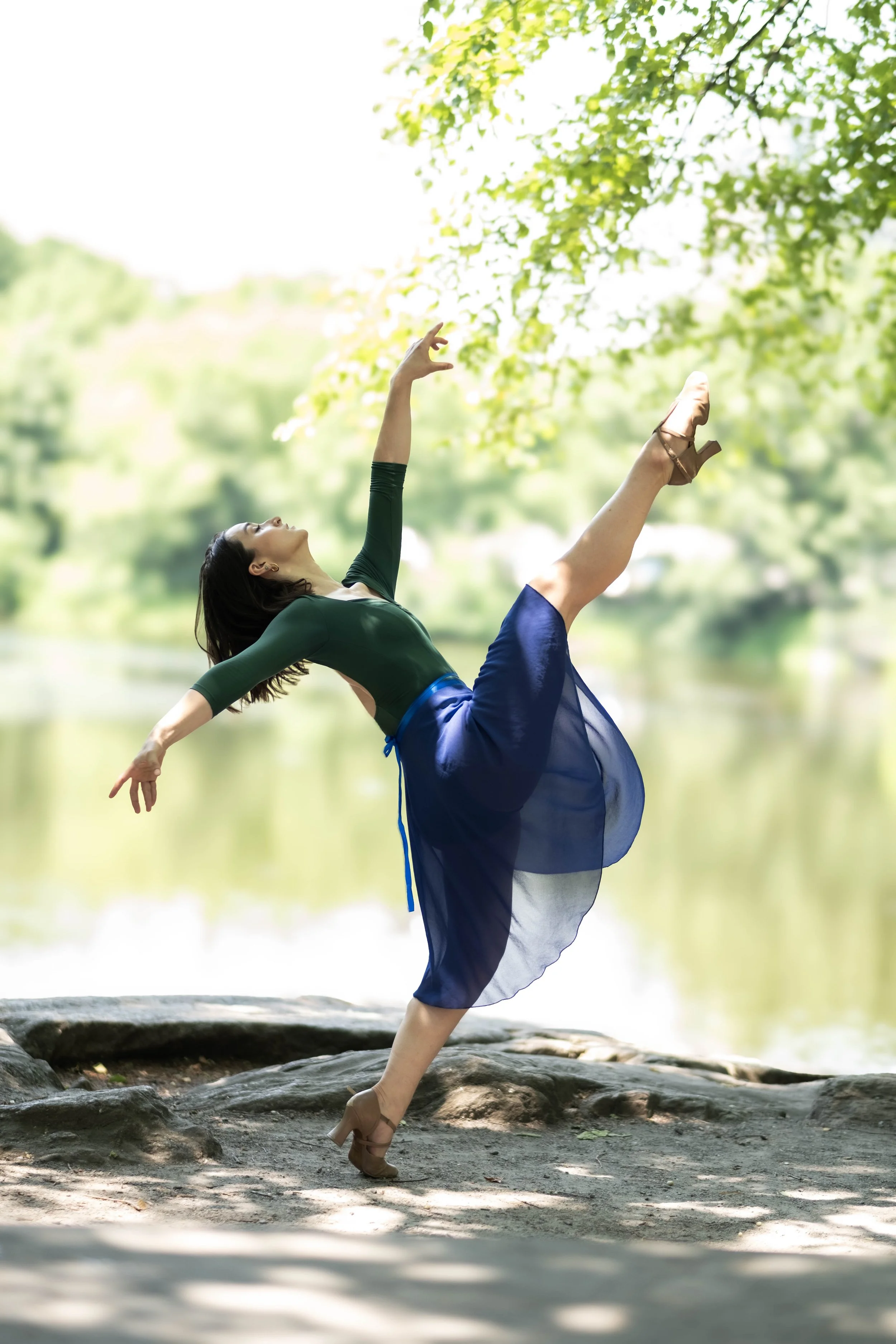 a woman practicing ballet outdoors near a body of water, standing on a rocky surface, wearing a green top, blue skirt, and brown heels, with one leg raised and arms extended gracefully