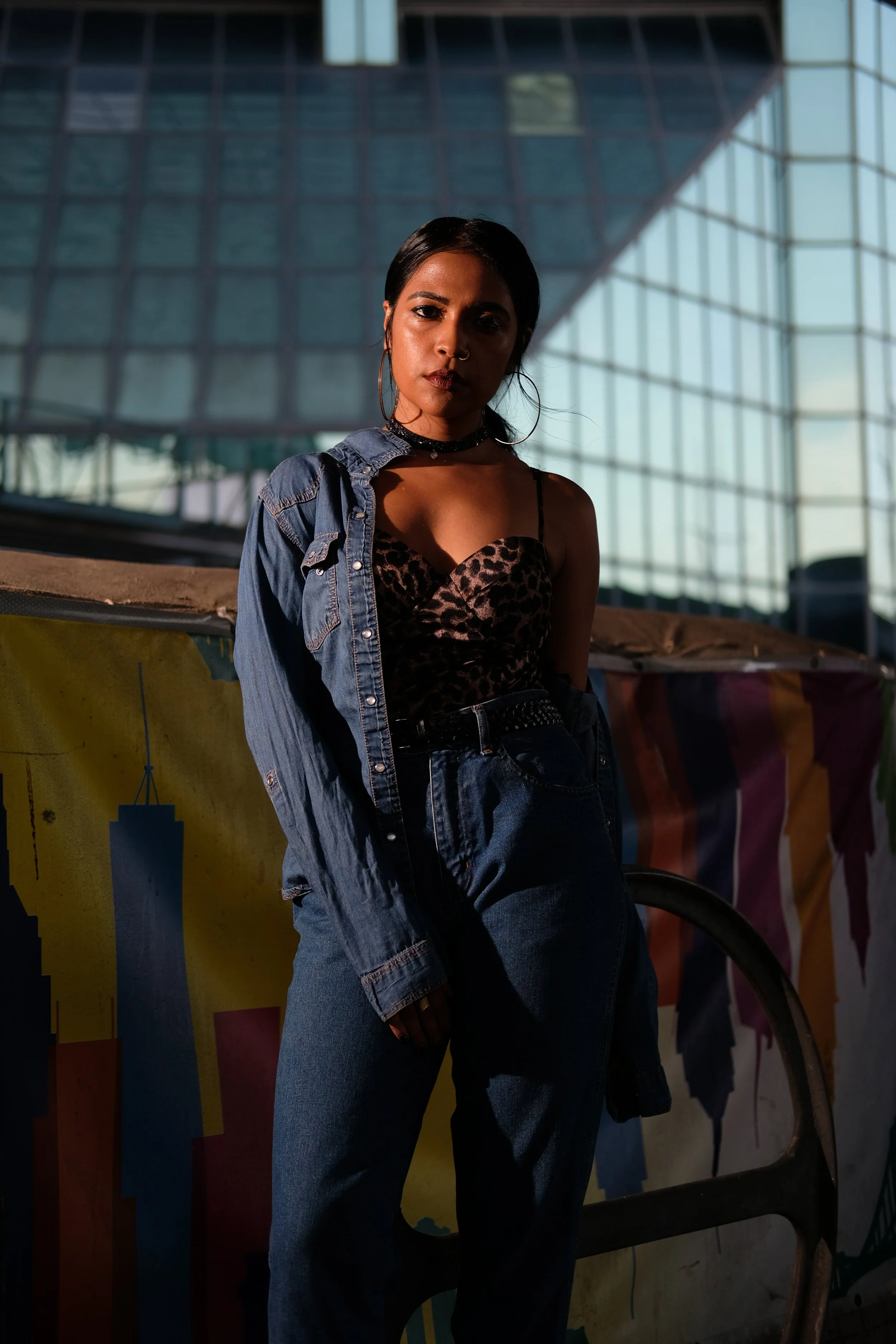 A woman with dark hair and dark clothing standing outdoors in front of colorful banners and a modern glass building.